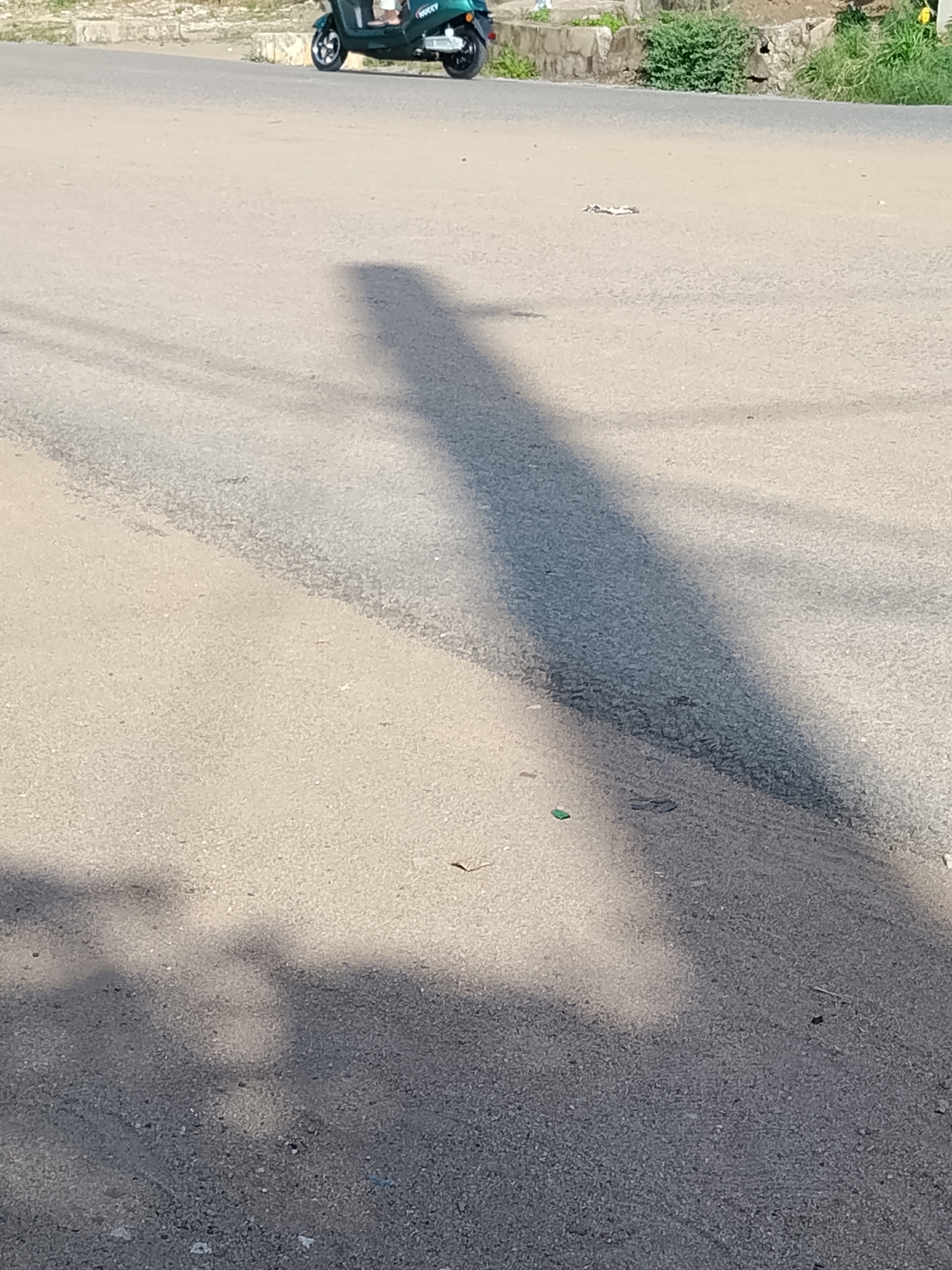 The image shows a shadow of a person on a paved road. In the background, a green scooter is parked. The scene appears to be in a residential area, possibly Wauro Leggal, Nigeria, with a stone wall and vegetation visible along the roadside. The lighting suggests it is daytime, with the sun casting a long shadow across the road. There are small pieces of debris scattered on the road surface.
