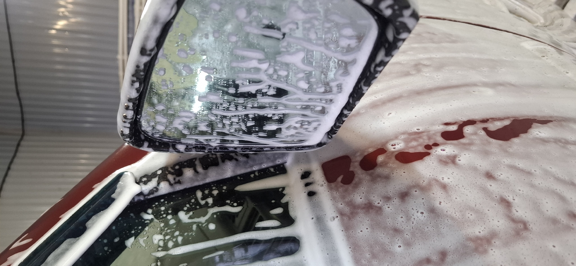 A close-up shot of a car being washed. Foam covers the car's body, side mirror, and window. The car's color appears to be red. The location is unspecified, and there's no indication of people or actions beyond the car wash itself. The environment seems to be an indoor or covered area, suggested by the overhead structure in the reflection. The only object identified is the car being cleaned. There is no verifiable location information.