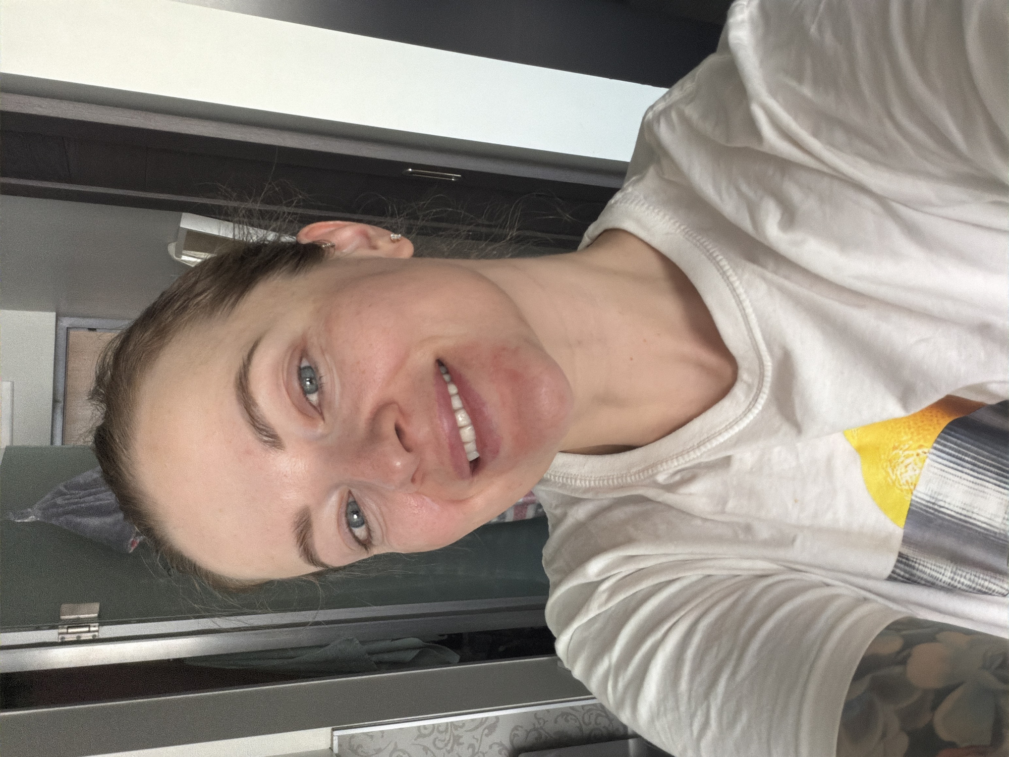A young woman with fair skin, blue eyes, and light brown hair pulled back is smiling at the camera. She has some redness on her cheeks and is wearing a white t-shirt with a yellow design on the sleeve. The background consists of a dark cabinet with a white top, possibly indicating an indoor setting like a bathroom or bedroom. Part of a window or mirror can be seen on the left side, along with a glimpse of a patterned object, possibly fabric or wallpaper. The presence of the cabinet suggests this image was likely taken indoors and may even be in a domestic setting, such as a residence in Moscow, Russia.
