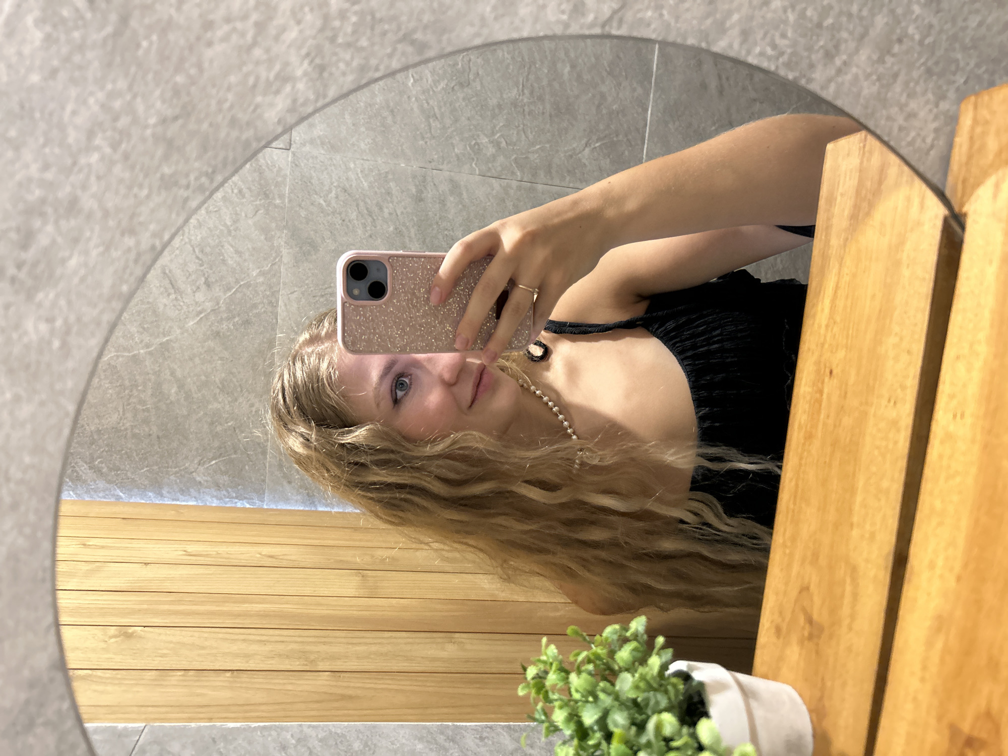 A person with long, wavy blonde hair and light-colored eyes is captured in a mirror selfie. They are wearing a black top or dress and a pearl necklace. A simple gold ring adorns the ring finger of their right hand, which is holding a smartphone. The phone features a distinctive pink, glittery case and a triple camera setup. The person has a gentle, pleasant expression with a slight smile.
The scene, reflected in the circular mirror, depicts a modern indoor space, likely a bathroom or dressing area. The background includes light grey, textured tiled walls. Below the tiled section, there are light-colored wooden panels arranged horizontally, suggesting a vanity or counter area. In the lower right portion of the reflection, a small potted plant with green foliage in a white pot adds a touch of greenery. The lighting appears bright and even, illuminating the scene. The image itself is rotated 90 degrees counter-clockwise. There is no visible text in the image.
