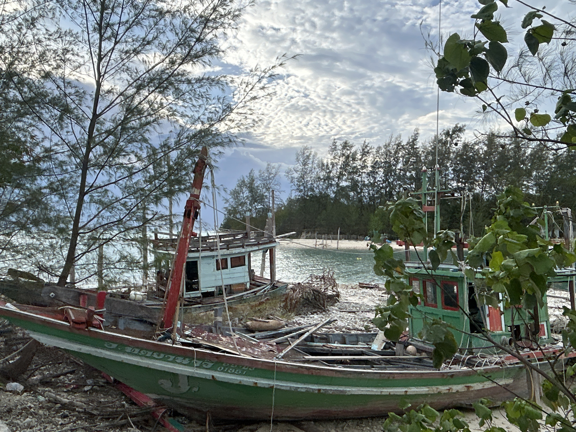 The image depicts a beach scene in ตำบลเกาะพะงัน, Thailand, featuring several beached and dilapidated fishing boats. In the foreground, a large green and red wooden boat rests on a pebble-strewn shore, its hull weathered and showing signs of disrepair, with visible debris within its structure. Behind it, another boat with a light blue cabin and a red mast is similarly beached. The background reveals a calm body of water with a sandy beach and a dense line of trees along the horizon. The sky is partly cloudy with a mix of white and gray clouds. Lush green foliage from trees and branches partially obscures the view in the foreground and upper right of the image. No people are visible in the scene.
