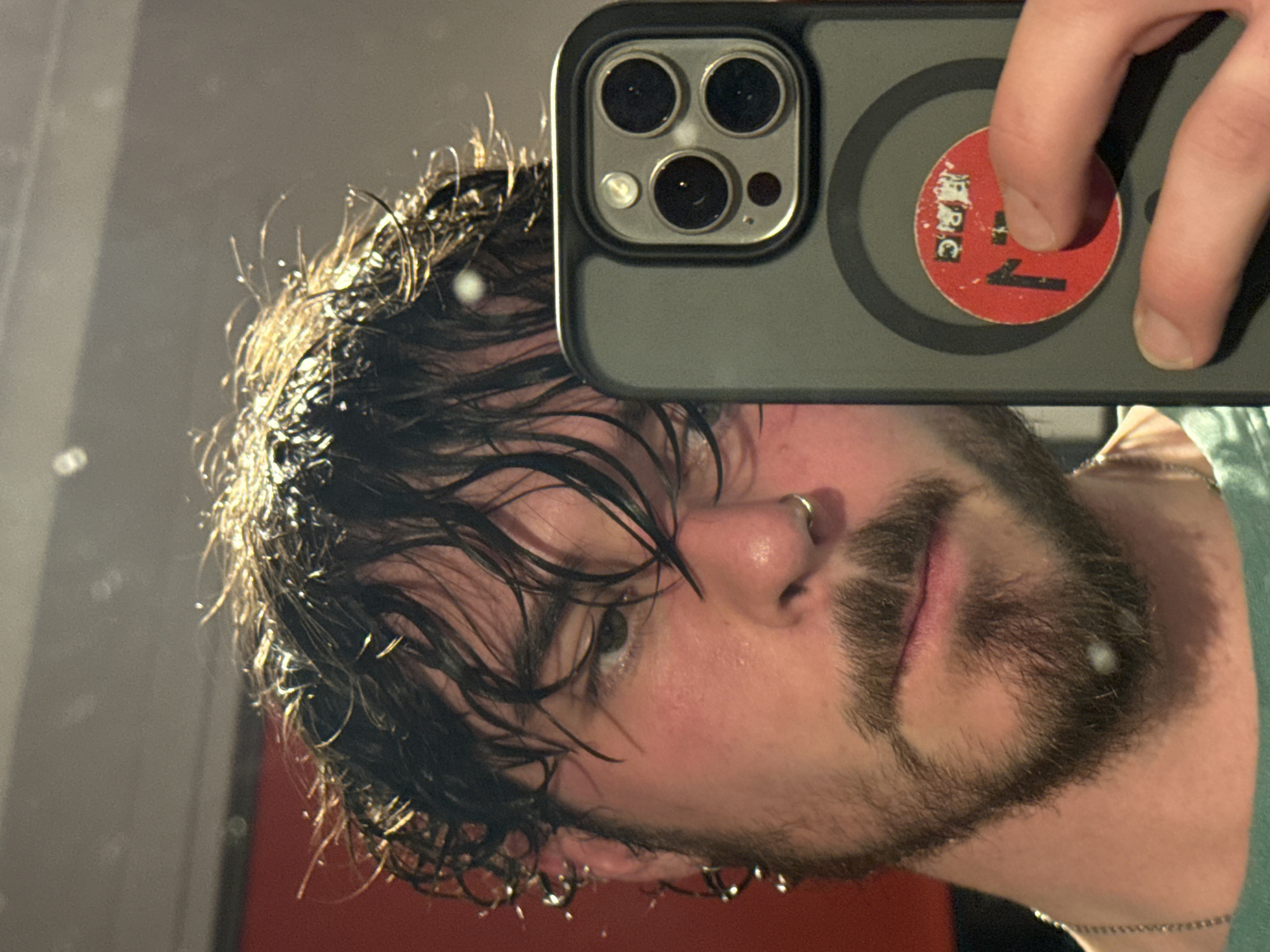 A man with wet, dark hair and a beard holds a phone up to a mirror, partially obscuring his face. He has a nose ring and appears to be in a bathroom, indicated by the reflective surface. The phone has a gray case with a circular red sticker with a "1" on it. The background suggests a red wall. The city of location is unknown.