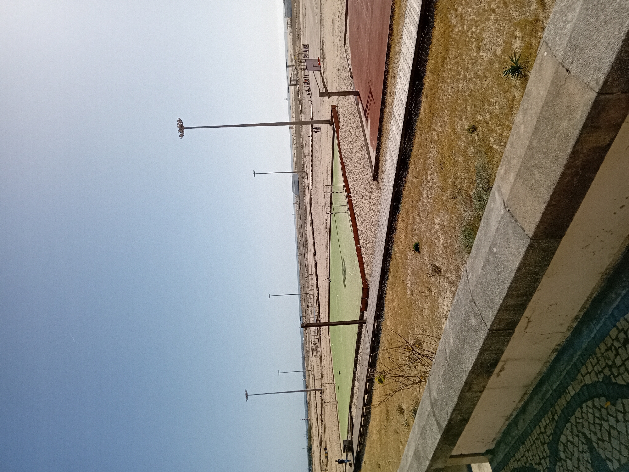 The image depicts a wide outdoor recreational area under a clear, bright sky, viewed from a slightly elevated perspective.
In the foreground, a robust light gray concrete railing or wall runs along the right side of the frame. Below this railing, a decorative section featuring dark blue and white mosaic tiling is visible. Beyond the railing lies a strip of dry, yellowish-brown grass, interspersed with a few small green plants.
The midground features a series of sports courts and sandy ground stretching into the distance. A wooden boardwalk or pathway separates the grassy area from the courts. The closest court is reddish-brown, equipped with a basketball hoop and backboard. Further along, a light green court, possibly for tennis or a similar sport, is visible. Several tall, slender light poles with multi-directional fixtures are spaced throughout the area, receding into the background. The ground between and beyond the courts is predominantly light-colored sand or dirt.
The background shows faint structures, possibly buildings or a monument, on the distant horizon, merging with the expansive sandy terrain. The sky, occupying the left half of the image, is a clear, light blue, with a subtle gradient to a lighter shade near the horizon. A faint white contrail from an airplane is visible in the sky.
The scene portrays a prepared recreational environment, but no specific activities are taking place. A single, small, indistinct human figure can be seen in the far distance near one of the light poles. The bright, clear sky suggests daytime and fair weather. The numerous light poles indicate the area is designed for use both day and night. The overall atmosphere is calm and largely deserted. No visible text is present in the image.