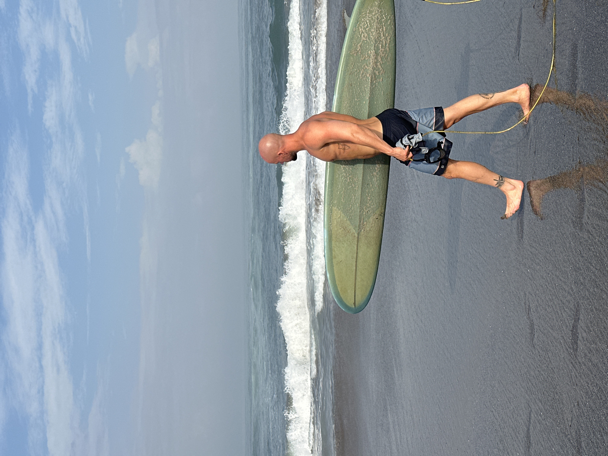 In North Kuta, Indonesia, a bald, fair-skinned man in dark blue and grey patterned swim trunks walks along a dark, wet sand beach, carrying a long, light green surfboard. The man is facing left, looking out at the ocean. He has tattoos on his left arm, right leg, and left foot. He is holding a grey towel and a black object in his left hand. A yellow surf leash trails behind him. The ocean in the background features moderate waves breaking, with white foam and spray. The sky above is light blue with scattered white clouds and a light haze near the horizon. The wet sand reflects the man and the surfboard.