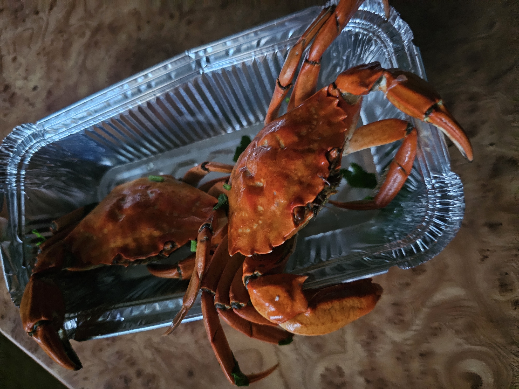 Two cooked crabs, an American blue crab and a mud crab, are displayed in a foil baking dish. The crabs are a vibrant orange-red color and their legs are spread out, showcasing their cooked state. The baking dish is placed on a wooden surface, possibly a table. The scene appears to be domestic, possibly a meal preparation or presentation. There is no information about the city of Skadovsk, Ukraine in the image.