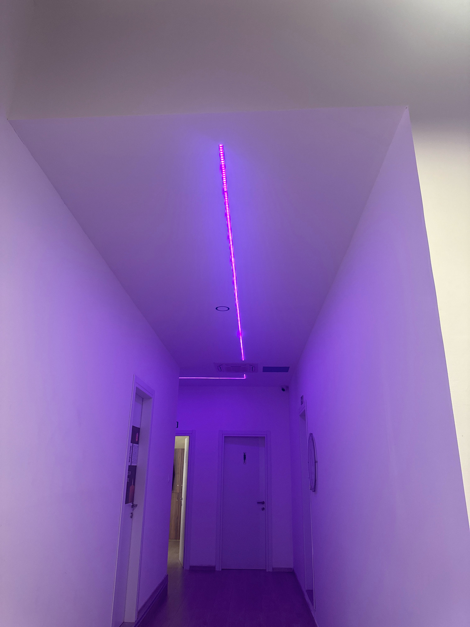 A long, narrow hallway is illuminated by purple LED lights. The hallway walls are white and appear to be painted. A strip of purple LED lights runs along the ceiling, casting a vibrant glow. There are several doorways visible along the hallway, suggesting this may be an office building or a residential complex. On the left side of the hallway, there is a white door with some posters or notices attached to it. Further down the hallway, on the right, there is a round mirror mounted on the wall. The floor of the hallway appears to be wooden. A security camera is visible on the ceiling in the distance. The overall impression is of a modern and well-lit space. The image is taken from a low angle, looking down the hallway towards the back. The location is in Germasogeia Municipality, Cyprus.
