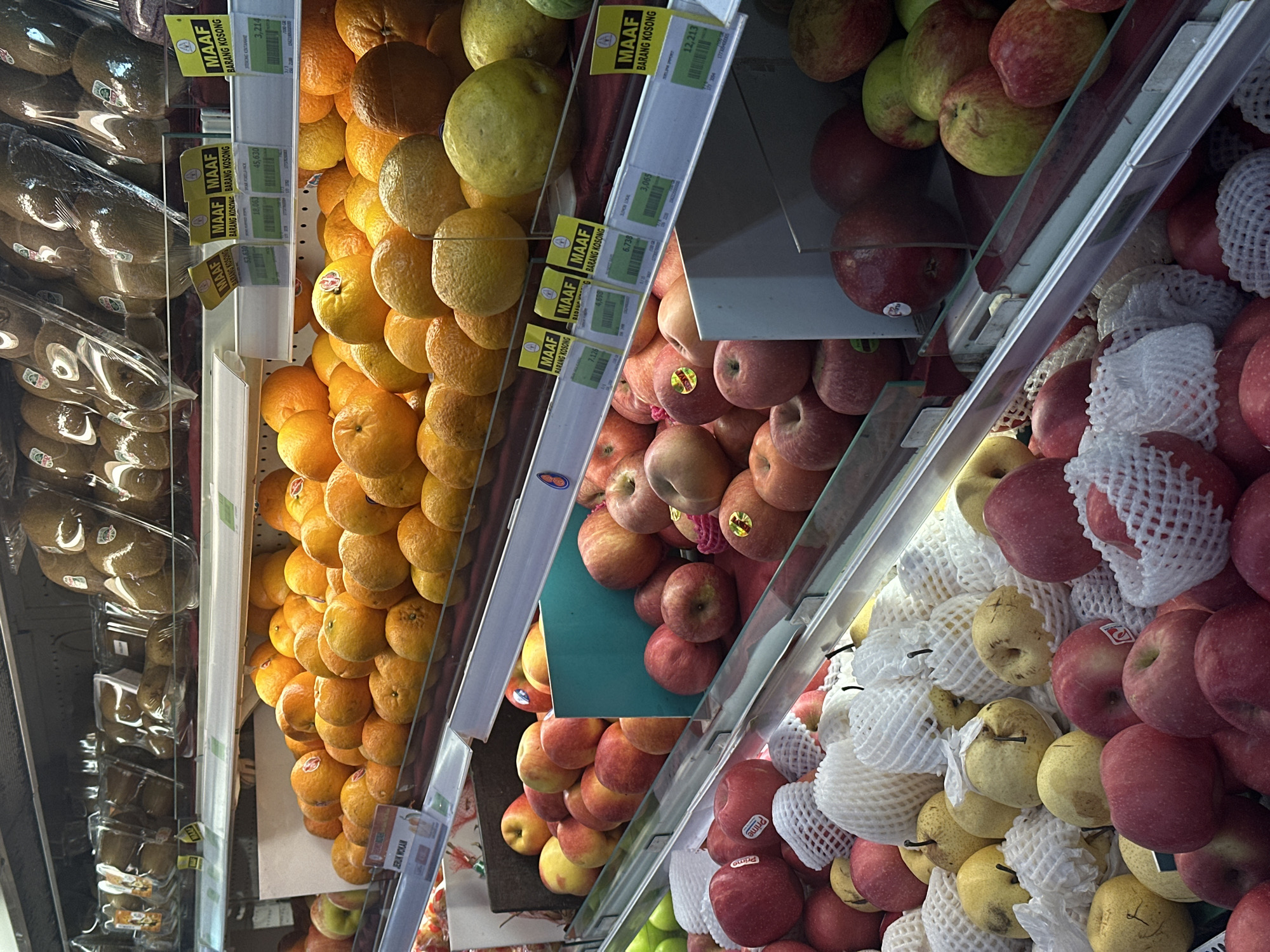 The image shows a brightly lit produce section of a supermarket, likely in North Kuta, Indonesia. The shelves are stocked with a variety of fruits. On the top shelf, there are multiple clear plastic packages containing kiwifruit, with some labels visible. The second shelf displays a large quantity of oranges, some loose and some in netting. Below this, there are displays of apples, both red and green varieties, some individually wrapped in protective netting. There are also some yellow-skinned pears visible. Price tags with Indonesian text are attached to the shelves in front of the fruit. The environment appears clean and organized, typical of a retail setting.