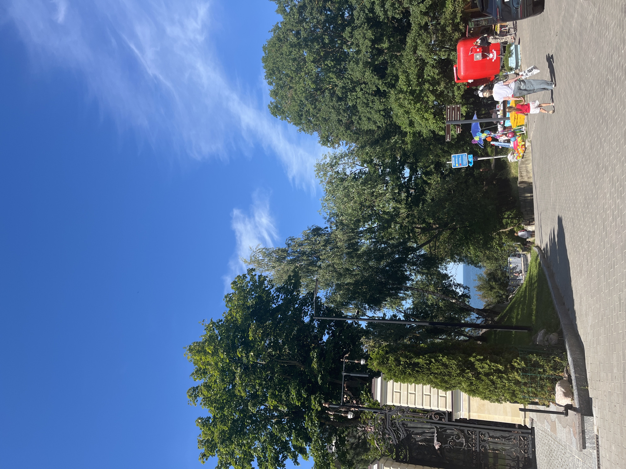 This is a street scene in Pionersky, Russia, on a sunny day with a clear blue sky and wispy clouds. The foreground shows a paved walkway made of grey rectangular bricks. To the left, a large ornate black metal gate stands next to a cream-colored stone pillar. A manicured evergreen hedge is planted in front of the pillar. Behind the gate and hedge, a very large, leafy green tree dominates the left side of the frame.
Further down the paved path, on the right side, two people are walking away from the camera: an adult wearing a white t-shirt and blue jeans, holding a shopping bag, and a young girl in a red shirt and shorts, holding the adult's hand. They are walking towards a red, somewhat retro-styled kiosk or food stand that has a blue umbrella and balloons nearby. Several other people are visible further in the background, near the kiosk and walking on another paved area. There are signs and more trees in the middle ground. In the distance, a body of water, likely the sea, is visible beyond a slope covered in green grass and trees. The overall atmosphere is casual and summery.