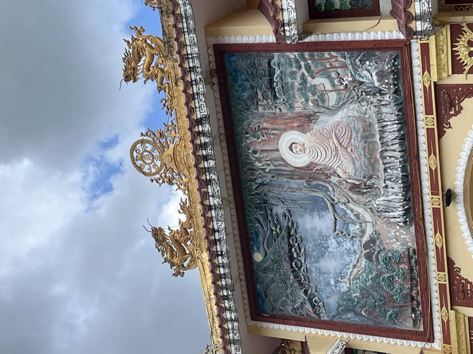 A segment of a temple or pagoda structure in Đăk Mar, Vietnam, is depicted under a partly cloudy sky. The upper portion of the image displays the building's roofline, adorned with intricate, golden, curved decorations. These include two prominent golden dragon statues positioned at the left and right ends of the visible roof section. Centrally located on the roofline is a golden Dharmachakra (Dharma wheel) symbol, flanked by additional golden ornate filigree. The roof features dark-tiled edges and a cream-colored frieze with a repeating pattern.
Below the roofline, a large, rectangular bas-relief sculpture occupies the central area of a cream-colored wall section. This relief features a depiction of Buddha meditating in the lotus position under a large tree, with a bright white halo behind his head. The landscape surrounding Buddha includes stylized mountains, coniferous trees, a body of water, and a full moon visible in the upper left portion of the relief. The relief is framed by a thick, dark red border. The building wall below the relief is a lighter cream color with additional red and golden architectural embellishments.