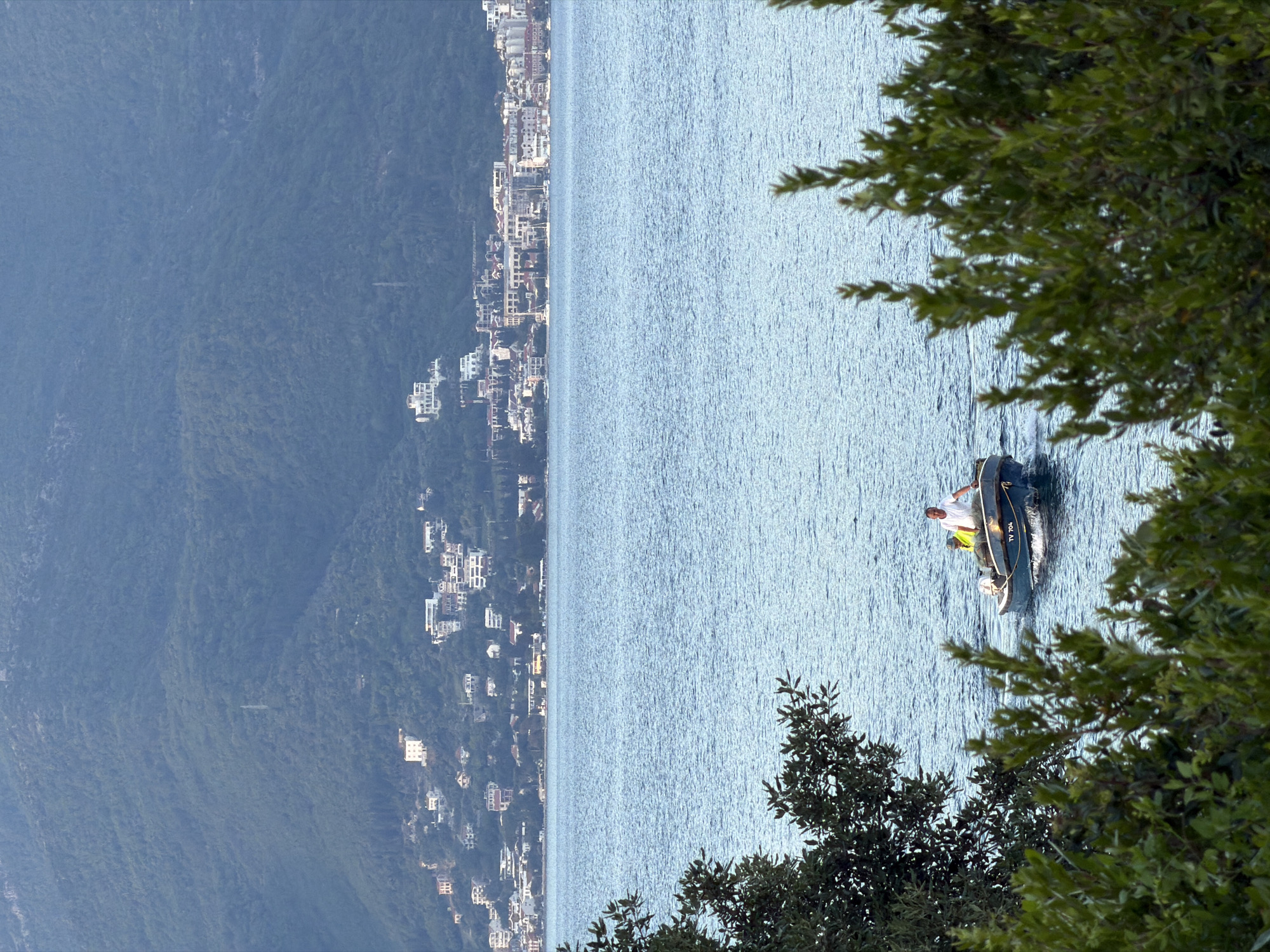 Viewed from land, the image shows a man in a small boat on the water, against a backdrop of the coastal city of Gošići, Montenegro, and a tree with green foliage framing the right side of the image. The man is wearing a white shirt and yellow vest and is in the boat, whose side is printed with "TV 704". The city is situated along the coast with buildings ascending along the hillside.
