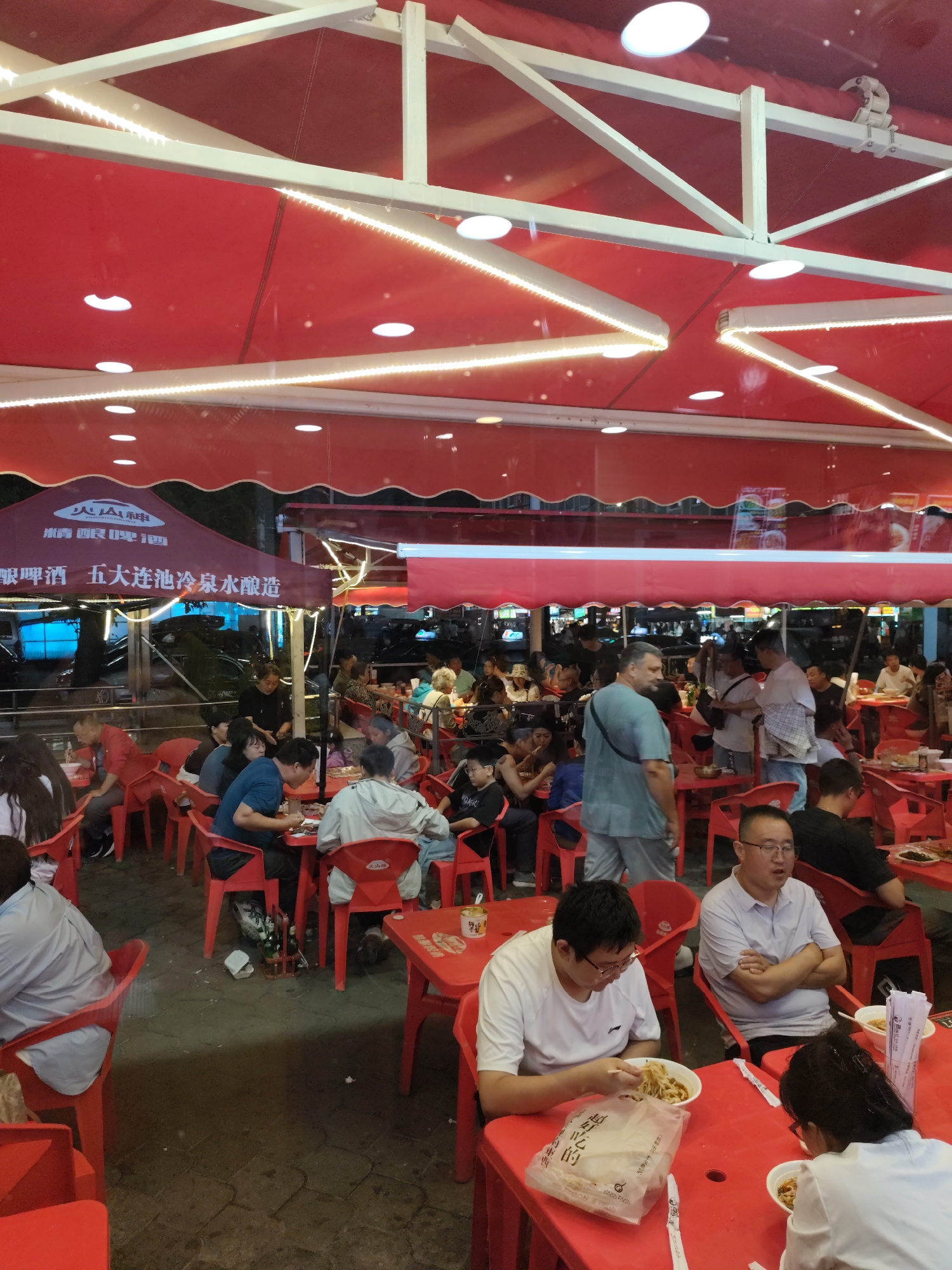 The image depicts a busy outdoor restaurant setting with numerous people seated at red tables and chairs, suggesting a lively dining atmosphere. The establishment appears to be covered by red awnings and canopies, with white structural beams and string lights overhead, indicating an evening or dimly lit environment. The patrons are diverse, with individuals and groups engaged in eating, conversing, and interacting. Some are actively consuming food, while others appear to be waiting or socializing.
The restaurant is likely a casual dining venue, possibly serving local cuisine, given the presence of food containers and bowls on the tables. The ground surface is paved with stone or brick patterns. The backdrop shows glimpses of vehicles and other structures, suggesting it is located within a city. The specific location of Aihui, China, is mentioned, implying that this scene is within that region. The overall impression is one of a bustling community gathering place focused on food and social interaction.