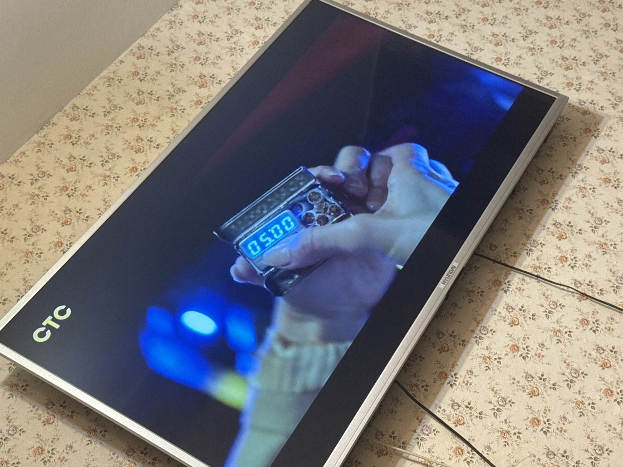 A television screen, displaying an image of a hand holding a cigarette case with a digital display showing "05:00", sits on a surface covered with a floral patterned cloth. The screen also shows part of a figure in the background illuminated by blue and green lights. The "CTC" logo is visible in the lower left corner of the screen. The television is branded "HYUNDAI" on the right side. The location is unknown in the city of Mytishchi, Russia.