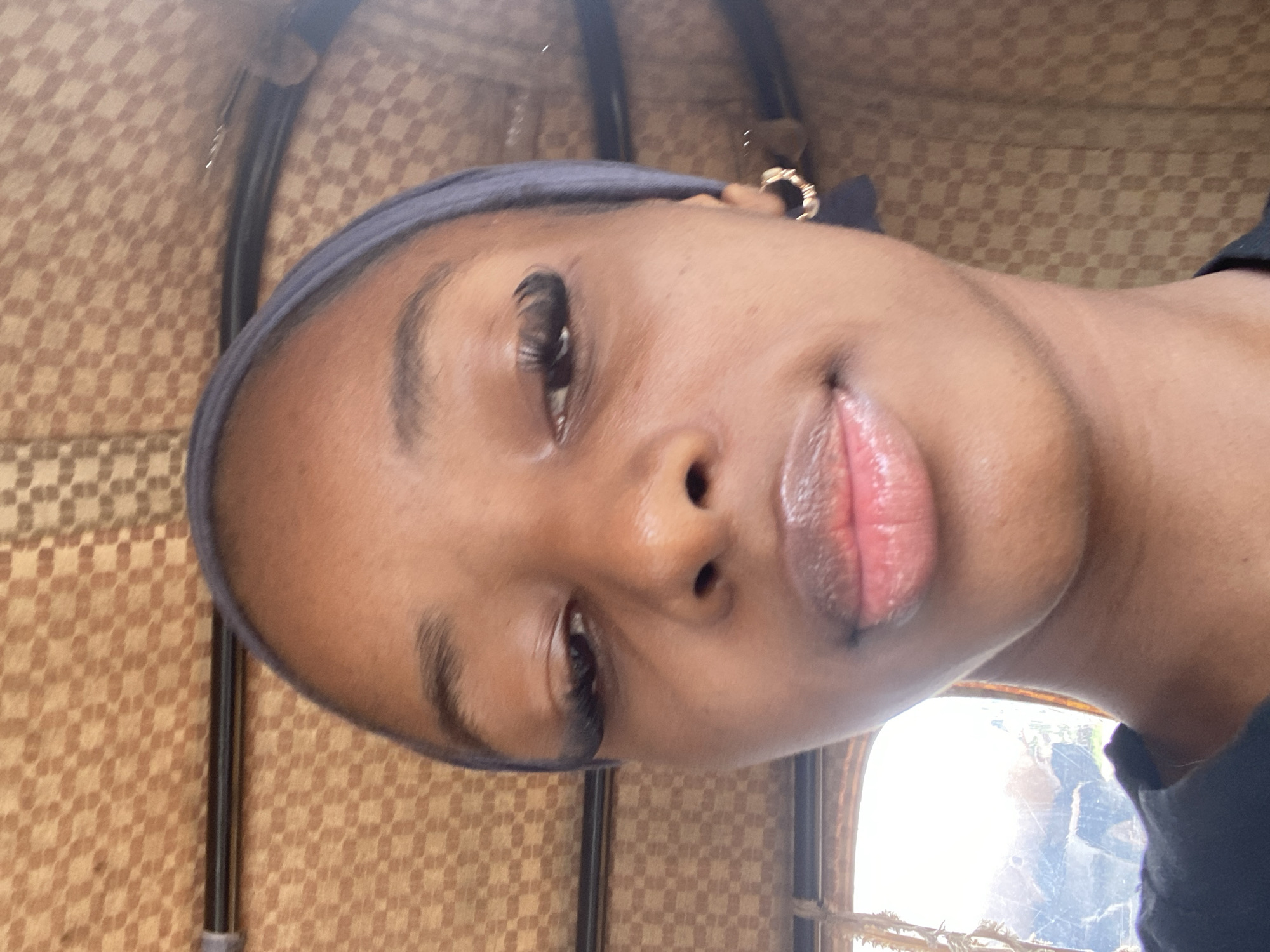 The image is a close-up, vertically oriented selfie of a young Black person. The subject is wearing a dark blue headscarf and a gold hoop earring in their left ear. Their eyes are open and looking towards the camera, with noticeable eyelashes. Their lips are slightly parted and appear to have a glossy finish.
The background is out of focus but shows a pattern of woven material, possibly upholstery or a wall covering, in shades of brown and beige. There are also dark vertical elements that could be part of a vehicle interior. To the right, a bright window with an arched top is visible, suggesting an indoor setting with natural light coming from outside. The lighting implies it could be daytime.
The overall impression is of a casual, personal photograph, possibly taken while traveling in a vehicle. No text is visible in the image.