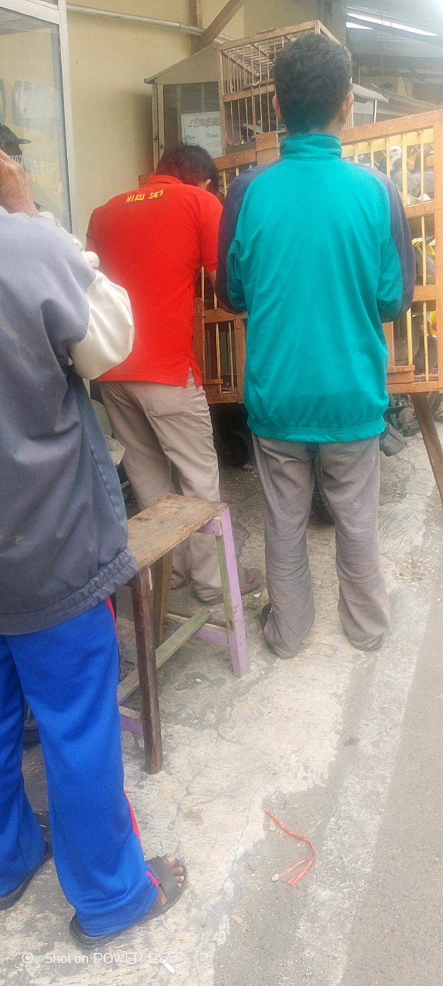 Three individuals are present in the outdoor scene. The person on the far left is wearing a dark blue jacket over a light-colored garment and blue pants. They are partially obscured, with only their torso and legs visible. In the center, a person in a bright red shirt with yellow lettering that reads "NIKU SET" and light-colored pants is facing away from the camera. To the right, another individual is wearing a teal jacket and gray pants, also facing away. They are standing near a wooden birdcage on a cart. In the foreground, a rustic wooden stool with purple legs is positioned on a concrete surface. The ground appears wet in places and is littered with debris. A thin red string with two ends is also visible on the ground. The overall setting suggests an urban environment, possibly near a market or a street vendor.