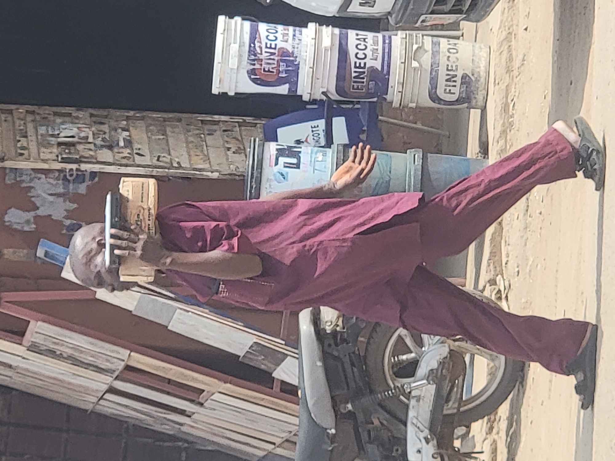 In Maiburiji, Nigeria, an elderly bald man dressed in a burgundy-colored traditional robe and pants is standing next to a parked motorcycle. He is holding a cell phone to his ear and speaking. Adjacent to him, stacks of wooden planks are visible along with several buckets of FINECOAT acrylic emulsion paint. The setting appears to be an outdoor area, possibly a roadside or a market location.