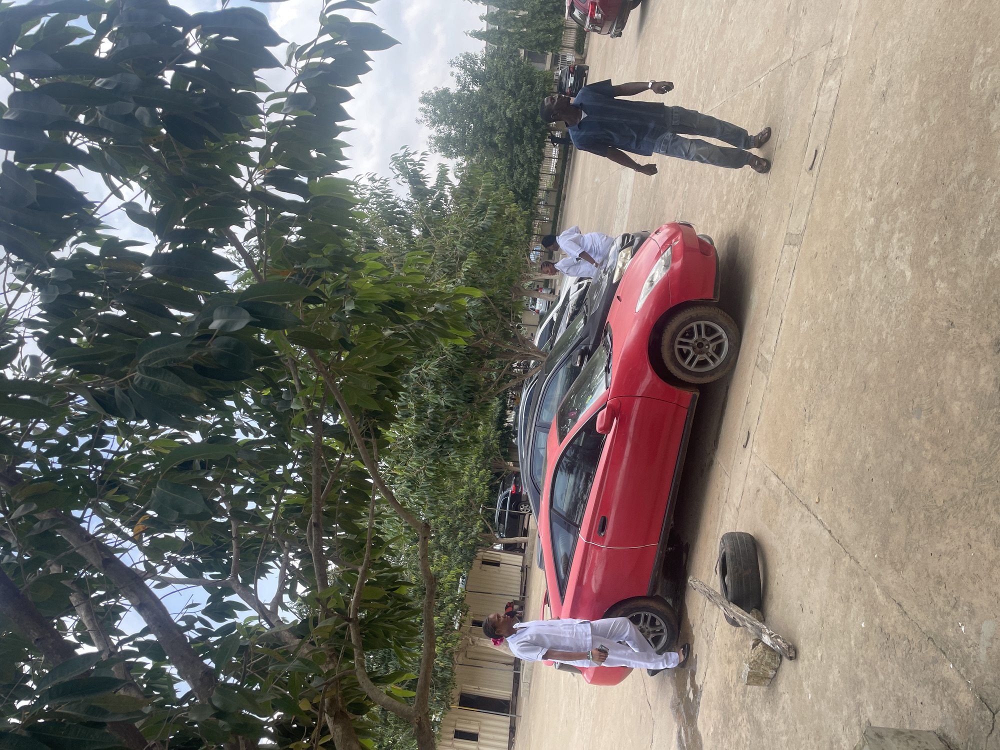 The image depicts an outdoor scene in Abuja, Nigeria, featuring several parked vehicles and individuals in what appears to be a parking lot or an open compound. Dominating the foreground is a bright red sports car, a Toyota Celica, positioned at an angle. To its left, a person dressed in white medical attire, possibly a nurse or doctor, stands next to the car, holding a mobile phone. Behind the red car, other vehicles are visible, including a black SUV and a darker sedan. In the mid-ground, another individual, a man wearing a blue patterned shirt and jeans, walks away from the camera towards the right. In the background, several long, rectangular structures, potentially modular buildings or shipping containers converted into rooms, are lined up. These structures are situated behind the parked cars, with trees providing shade over the scene. The ground is paved with concrete, showing some cracks and a discarded tire and piece of wood in the lower left portion of the image. The sky is overcast, suggesting a cloudy day. The overall impression is one of a functional space, perhaps associated with a medical facility, school, or business premises, given the presence of the individuals in medical attire and the utilitarian buildings.