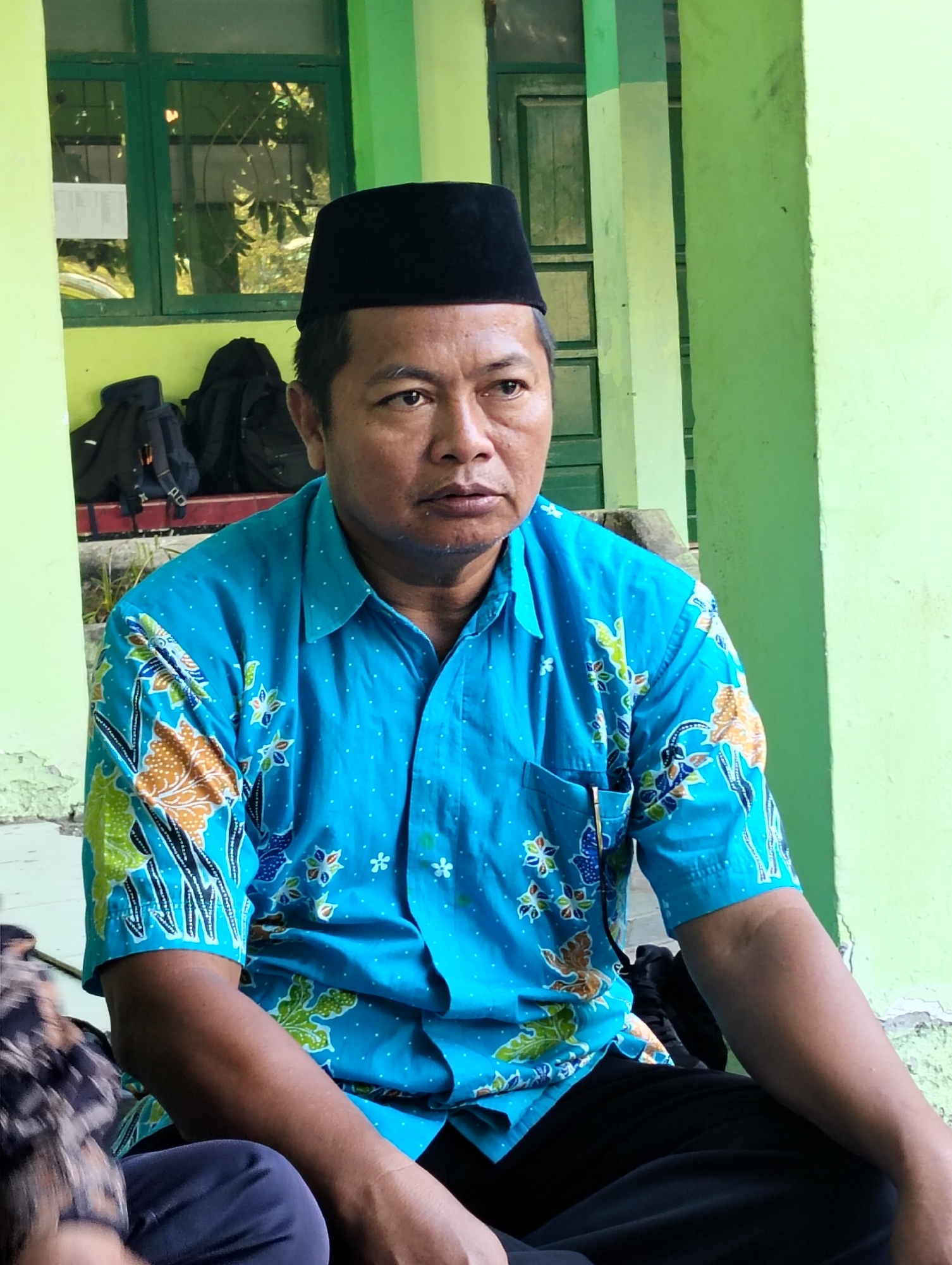 A middle-aged man with dark hair and a black peci hat is seated outdoors. He is wearing a bright blue, short-sleeved batik shirt with white polka dots and a floral pattern in shades of orange, green, and blue. He is looking off to the right with a neutral expression. Behind him, a light green wall with some peeling paint is visible, along with a green-framed window. Two black backpacks are placed on a raised surface to the left of the man. The overall impression is a candid, everyday scene possibly in a public area of Dagan, Indonesia.