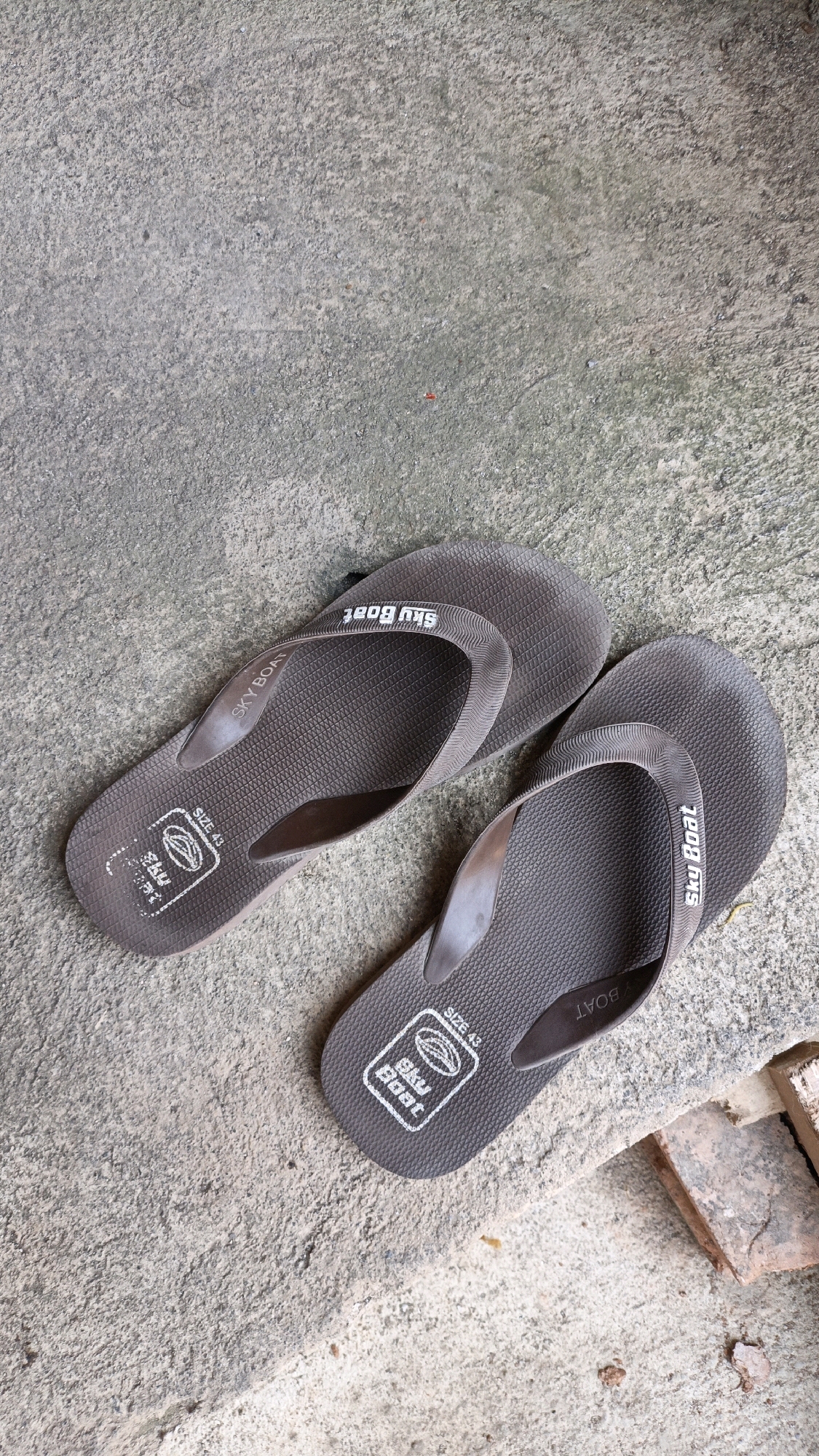 Two brown flip-flops are lying on a textured concrete surface. The flip-flops are positioned diagonally, with the left one slightly above and to the left of the right one. Both flip-flops display the brand name "SKY BOAT" in white lettering on the straps and on the footbed. The footbed also shows a size indication of "SIZE 43" along with a logo. The concrete surface appears weathered and uneven. In the lower right corner of the image, a few bricks are partially visible, suggesting an outdoor or construction-adjacent environment. This scene is consistent with an outdoor area in Cikal, Indonesia, given the commonality of flip-flops and the typical appearance of outdoor surfaces and construction materials in the region.