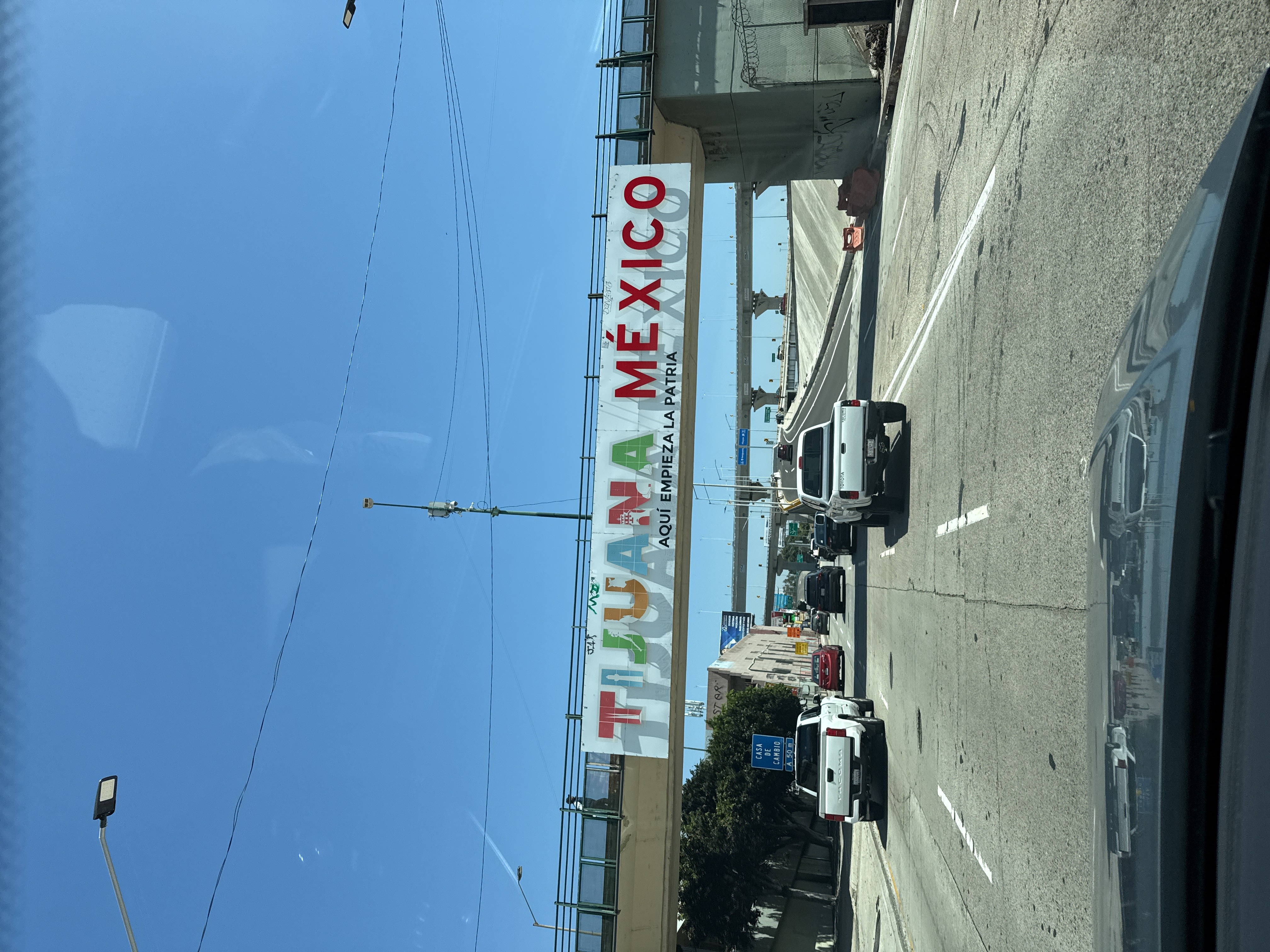 The image shows a street in Tijuana, Mexico, beneath a bridge displaying a sign that reads "TIJUANA MEXICO AQUI EMPIEZA LA PATRIA." Several vehicles are visible on the road, including two white pickup trucks in the foreground. The sky is clear and blue. The photo appears to have been taken from inside a vehicle.