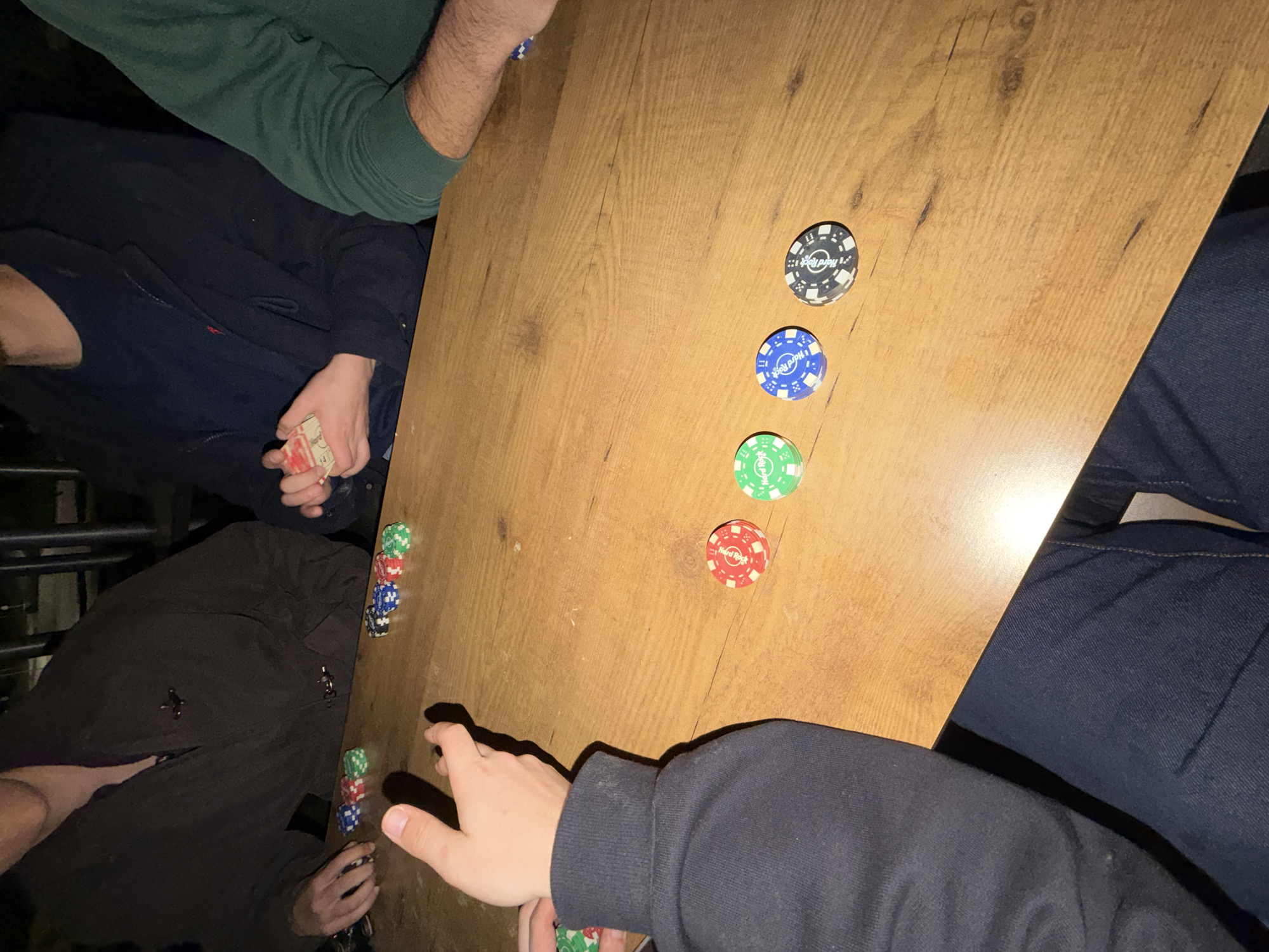 A top-down shot shows people playing poker on a wooden table. Poker chips of various colors (black, blue, green, red) are scattered across the table. The players are wearing casual clothing, including a green sweater, a black jacket, and jeans. One player is holding a stack of cards. The location appears to be indoors, possibly a private residence. The location of Vila Real, Portugal, is not apparent from the visual content of this image.