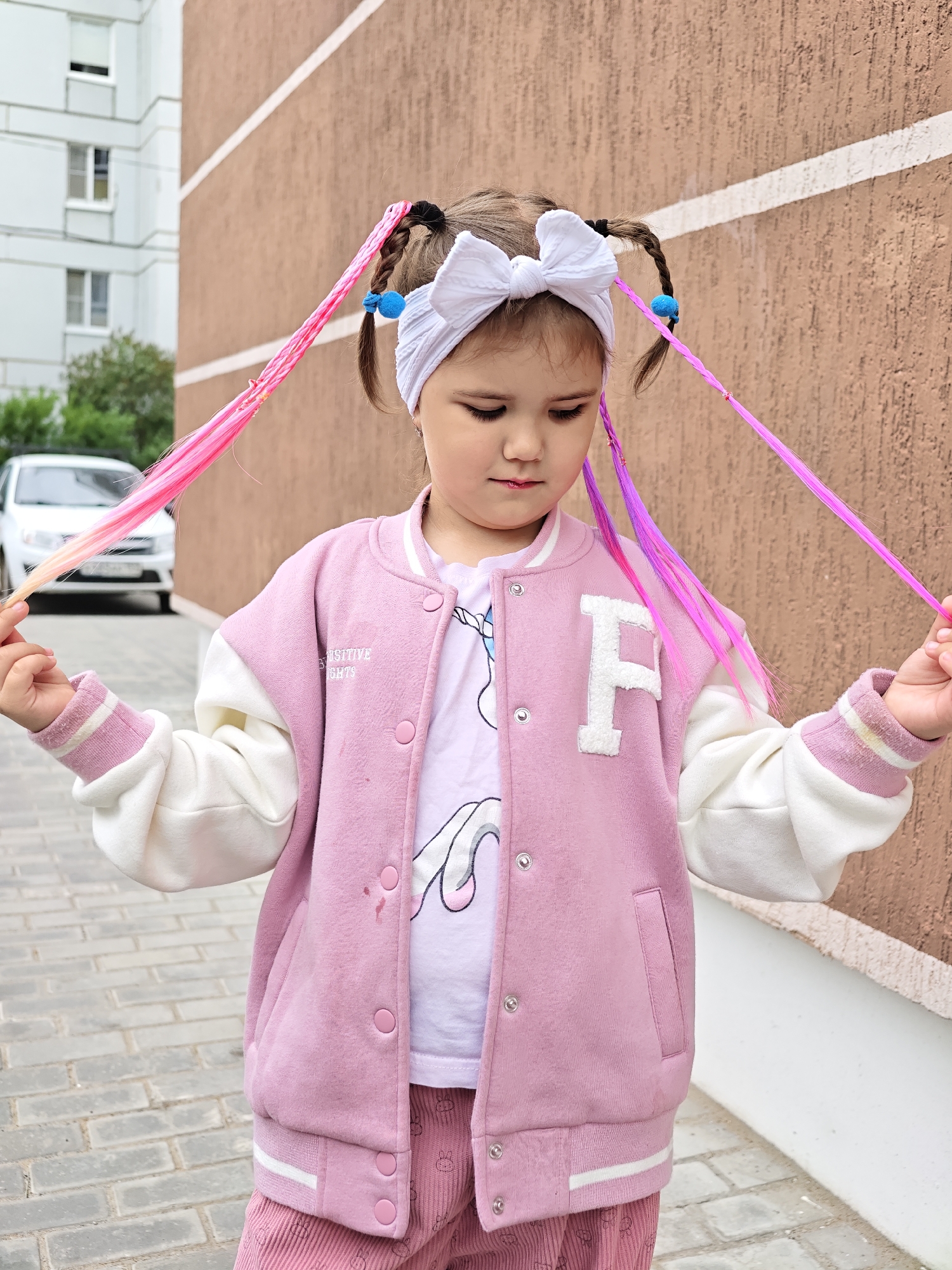 A young girl with braided pigtails adorned with pink and purple extensions stands outdoors, looking down with a neutral expression. She is wearing a pink and white bomber jacket over a t-shirt with a unicorn graphic and pink corduroy pants. A white bow is tied on top of her head. The background features a textured brown wall with white horizontal stripes, a brick walkway, and a white car parked on the left. The scene appears to be in a residential area, possibly in Chaikovsky, Russia.