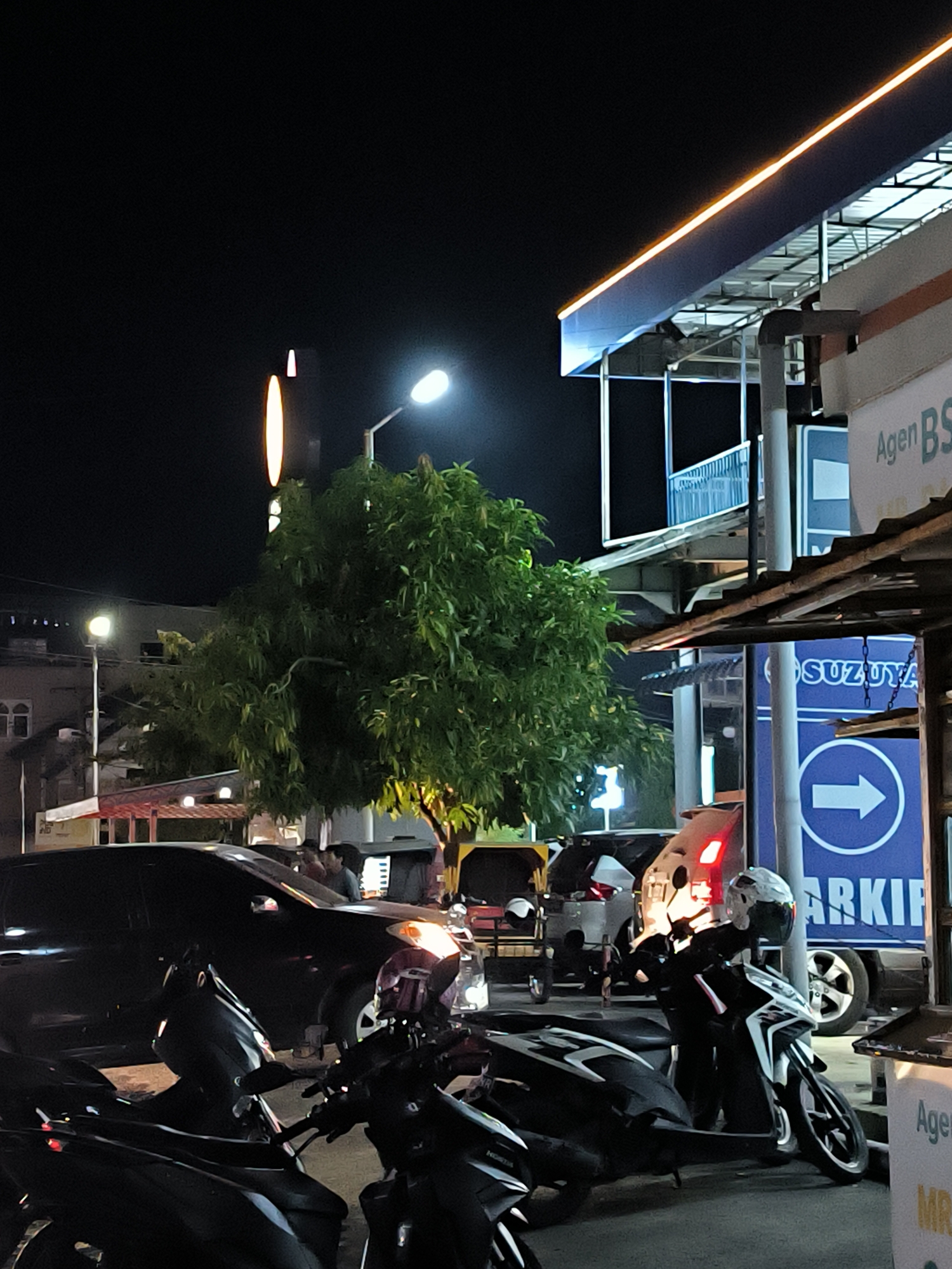 The image depicts a nighttime scene in Lhokseumawe, Indonesia. Several motorbikes are parked in the foreground. Behind them, cars are lined up on the street. A large tree is near the center of the frame, illuminated by a street lamp. Signage for "SUZUYA" and "AGEN BS" can be seen on the right side of the image, indicating a business or establishment. The upper portion of a building with a brightly lit ledge is visible in the upper right. In the distance, a tall, thin structure is illuminated by an orange light. The image is dimly lit, with several light sources creating a contrast between the dark sky and the illuminated objects in the foreground.