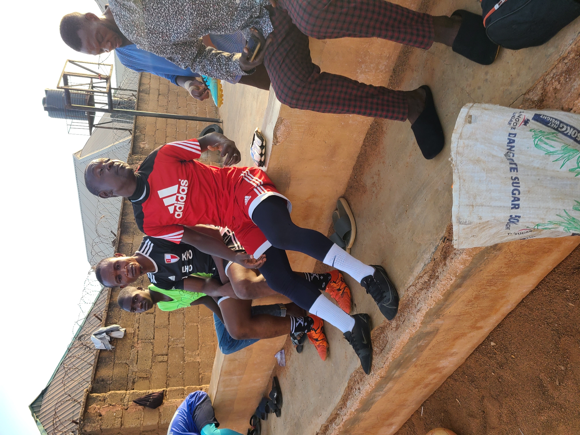 The photograph shows a group of men outdoors, likely in a semi-urban or rural setting. One man is wearing a red Adidas jersey and black leggings with white socks, while others are in athletic attire, suggesting they may be involved in sports or exercise. A bag of "DANGOTE SUGAR 50KG" is also in the image. The location is Unknown, Unknown.