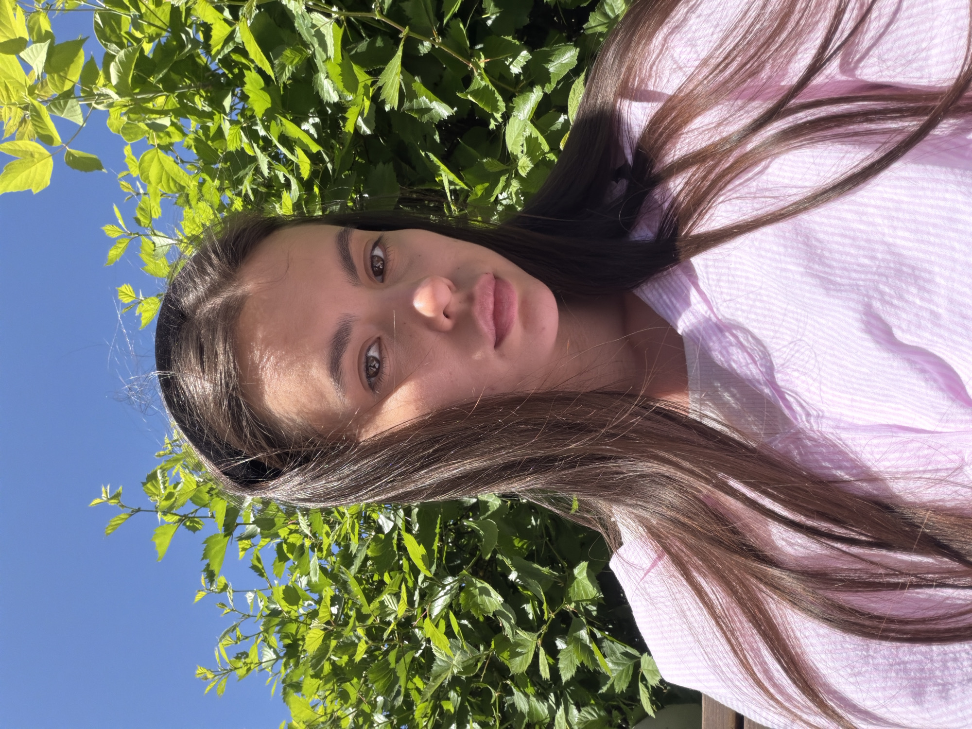 A young woman with long, dark hair is positioned in front of greenery, likely foliage from a tree. She is wearing a light pink striped shirt. The background includes a blue sky visible through the leaves. The specific location within Chaikovsky, Russia cannot be determined from the image alone. There is a single person present. She does not appear to be interacting with anything specific.