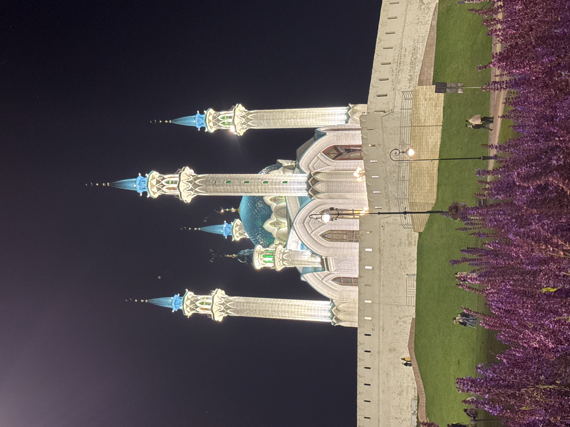 The image shows the Kul Sharif Mosque in Kazan, Russia, at night. The mosque is illuminated, with its white walls and blue-domed minarets standing out against the dark sky. A white stone wall with small openings surrounds the mosque, and a green grassy slope leads up to it. In the foreground, a cluster of purple flowers is in bloom. Several people are visible on the lawn and walkway in front of the mosque, some walking and others standing or sitting. The overall scene is serene and picturesque, highlighting the architectural beauty of the mosque and its surroundings.
