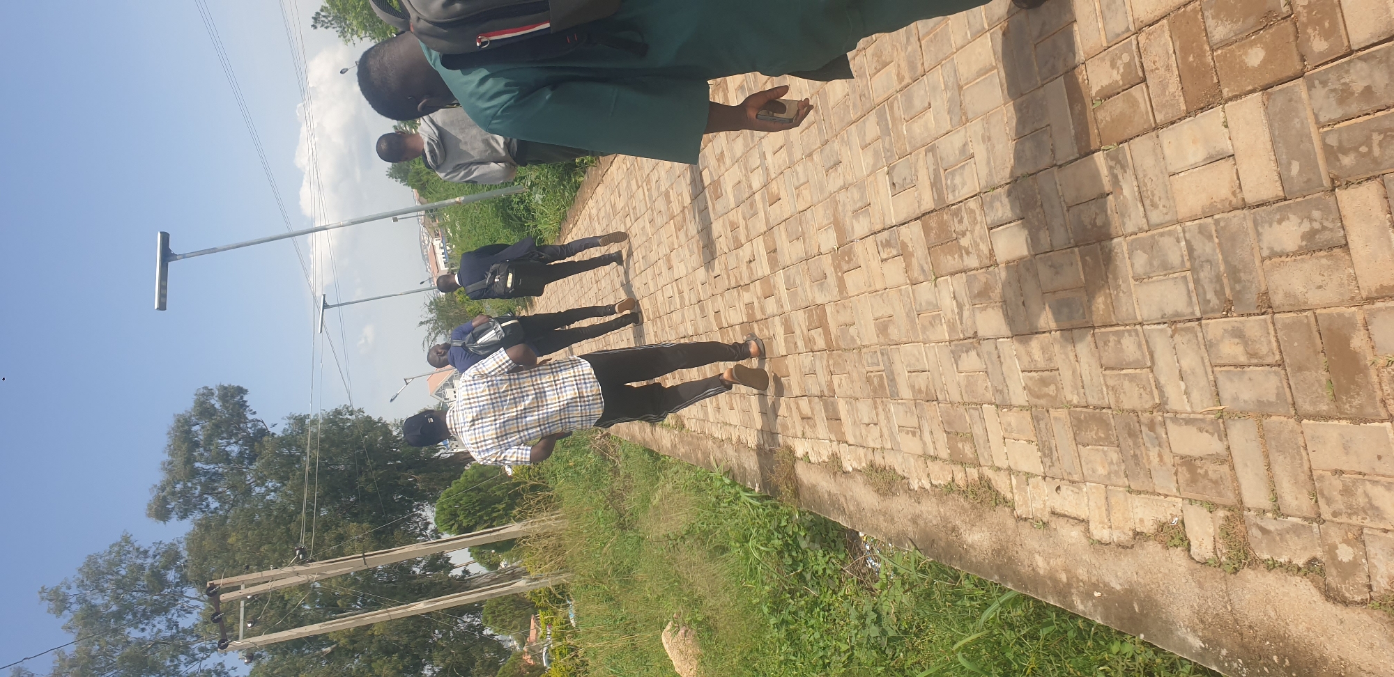 In Rafin Par, Nigeria, five individuals are observed walking along a paved walkway. The path is constructed of rectangular bricks laid in a herringbone pattern. To the left of the path, there is overgrown green grass and vegetation. In the background, trees with lush green foliage are visible, along with utility poles and wires. The sky is clear and blue with a few scattered clouds. The sun is bright, casting shadows from the people and other objects onto the walkway.
The individuals are walking away from the camera, moving towards the right side of the image. The person closest to the camera, on the right, is wearing a green robe and appears to be holding a cell phone. The person in front of them is wearing a grey hoodie. The third person from the right is wearing a dark suit and carrying a black backpack. The fourth person is wearing a blue shirt and also carrying a black backpack. The person furthest to the left is wearing a checkered shirt and black pants. All of them are walking in the same direction.
Two streetlights with solar panels are visible in the background, suggesting the presence of a well-maintained public area. The overall environment suggests an outdoor setting on a sunny day.
