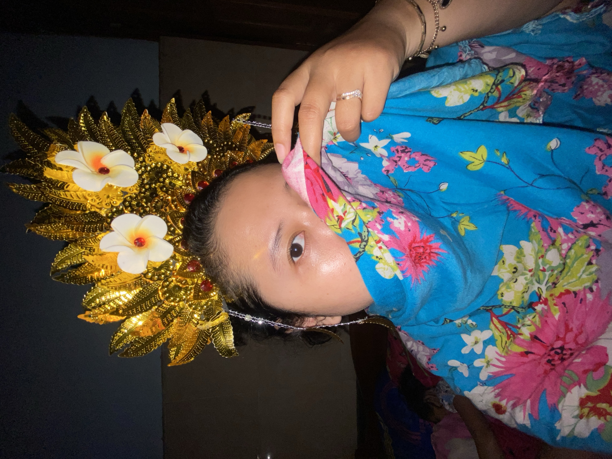 A close-up, dimly lit portrait depicts a female subject indoors, located in Gelam, Indonesia. The subject's face is partially obscured by a folded piece of blue fabric patterned with large pink, white, and green flowers, held by her visible right hand over her mouth and nose. Her right eye is visible and directed towards the camera. She is wearing an elaborate golden headpiece featuring multiple layered, leaf-like structures, three prominent white Plumeria-like flowers with orange-red centers, and red gem accents. A beaded ornament hangs from the headpiece near her right ear. Her visible right hand displays a two-band ring on the ring finger and multiple chain bracelets on the wrist. The background is dark and blurred, revealing indistinct shapes and colors consistent with soft furnishings in an indoor environment.
