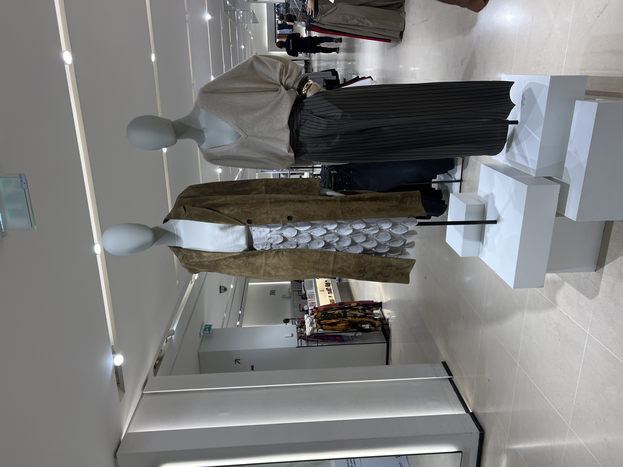 The image shows a retail store interior, likely a clothing boutique, in Nice, France. Two mannequins are prominently displayed in the foreground, showcasing outfits. The mannequin on the left wears a long, brown suede-like jacket over a white top and a white, scaled skirt. The mannequin on the right is dressed in a beige V-neck sweater and high-waisted, pinstriped gray pants. Both mannequins are elevated on white tiered platforms. In the background, a person in a dark uniform, possibly a security guard or employee, stands near a rack of clothing. The store environment features bright overhead lighting, tiled flooring, and various displays of merchandise. Signs are visible in the background, including a green exit sign with an arrow, and a sign that appears to read "ZARA". The overall scene depicts a typical retail setting with mannequins used to present fashion items.