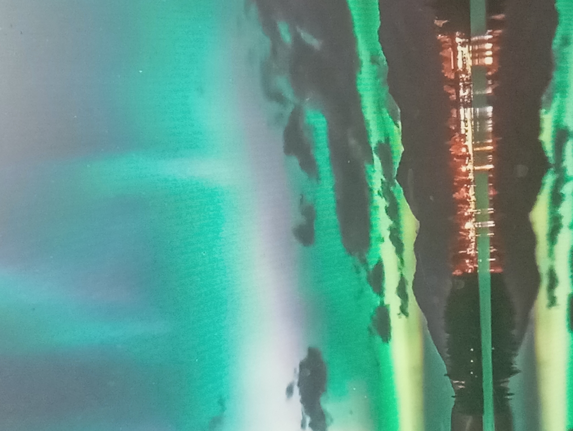 I am unable to identify the location as Karaganda, Kazakhstan from the given image. The image shows what appears to be a reflection in water. There are blurry green, blue, and white colors in the background. A dark shape is in the center, which may be trees or another structure reflected in the water. Some orange or yellow light is visible in the center as well.