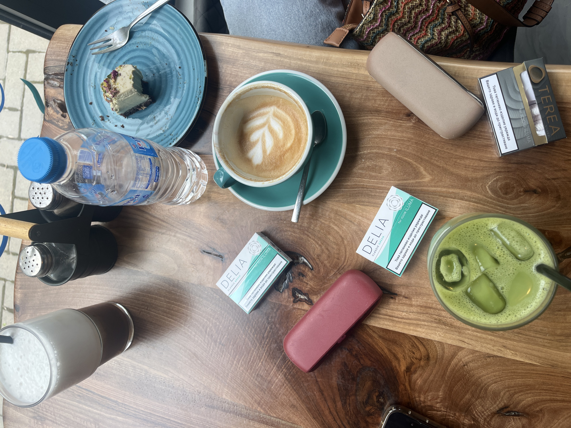 An overhead shot captures a wooden table laden with various items in what appears to be a café or similar establishment, situated in Plovdiv, Bulgaria, as indicated by the Cyrillic text. The table features a partially eaten slice of dessert, possibly pistachio cake with rose petals, on a textured blue plate with a fork. Next to it is a plastic water bottle with a blue cap. A cup of coffee with latte art sits in a teal saucer, accompanied by a spoon.
Other beverages include a tall glass with a dark liquid and a foamy white top, and another tall glass containing a vibrant green, ice-filled drink with a straw, likely a matcha latte. Salt and pepper shakers are also visible. Personal items scattered across the table include a beige rectangular case, a red rectangular case, and part of a colorful woven bag or purse. A corner of a smartphone is seen at the bottom right.
Three boxes of heated tobacco sticks for IQOS ILUMA are present: two "DELIA CLASSIC GREEN" and one "TEREA SILVER". All boxes display the warning in Bulgarian Cyrillic: "Това тютюнево изделие уврежда Вашето здраве и води до пристрастяване" (This tobacco product harms your health and causes addiction). The overall scene suggests a casual gathering or a relaxed break, enjoying drinks and a snack during daytime.