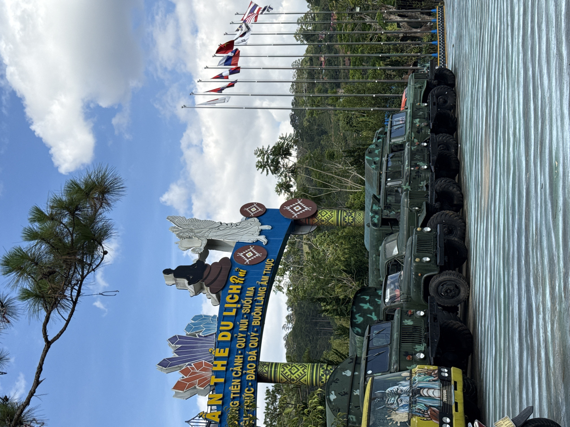 The image depicts a row of military trucks parked next to a sign for "An The Du Lich Rui," a tourist attraction in Păng Tiêng 1, Vietnam. The sign is colorful and decorated with various shapes and designs. Flags are also flying in the background. The weather appears sunny with scattered clouds. The surrounding environment includes trees and some hilly terrain.