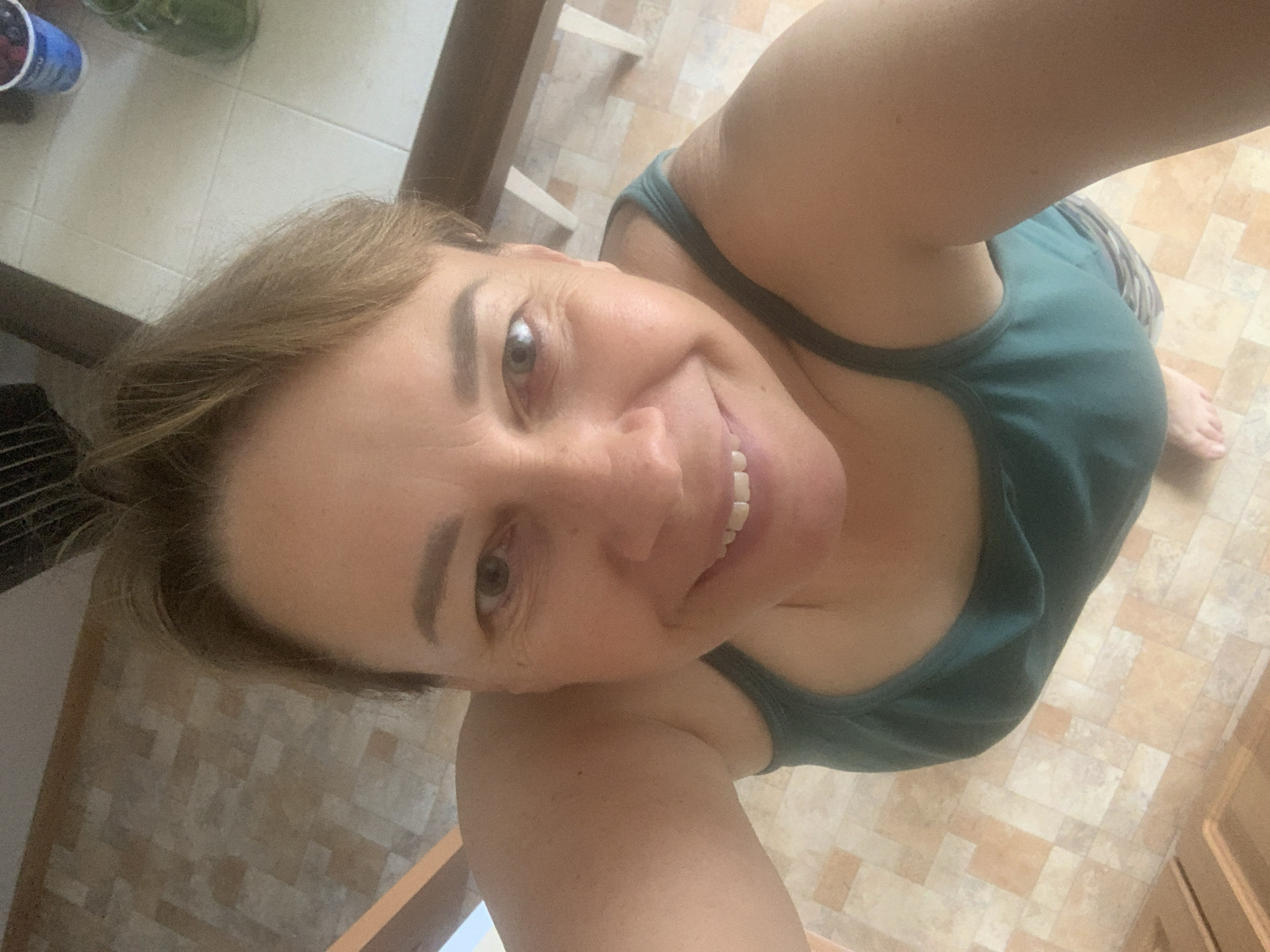 The image shows a selfie taken from a high angle, looking down at a woman. The woman has medium-length brown hair and is smiling at the camera. She is wearing a teal tank top. The background appears to be an indoor setting with tiled flooring. There are hints of wooden furniture or structures in the background. There are no discernible features that would suggest the location is the city of Kapyĺ, Belarus.