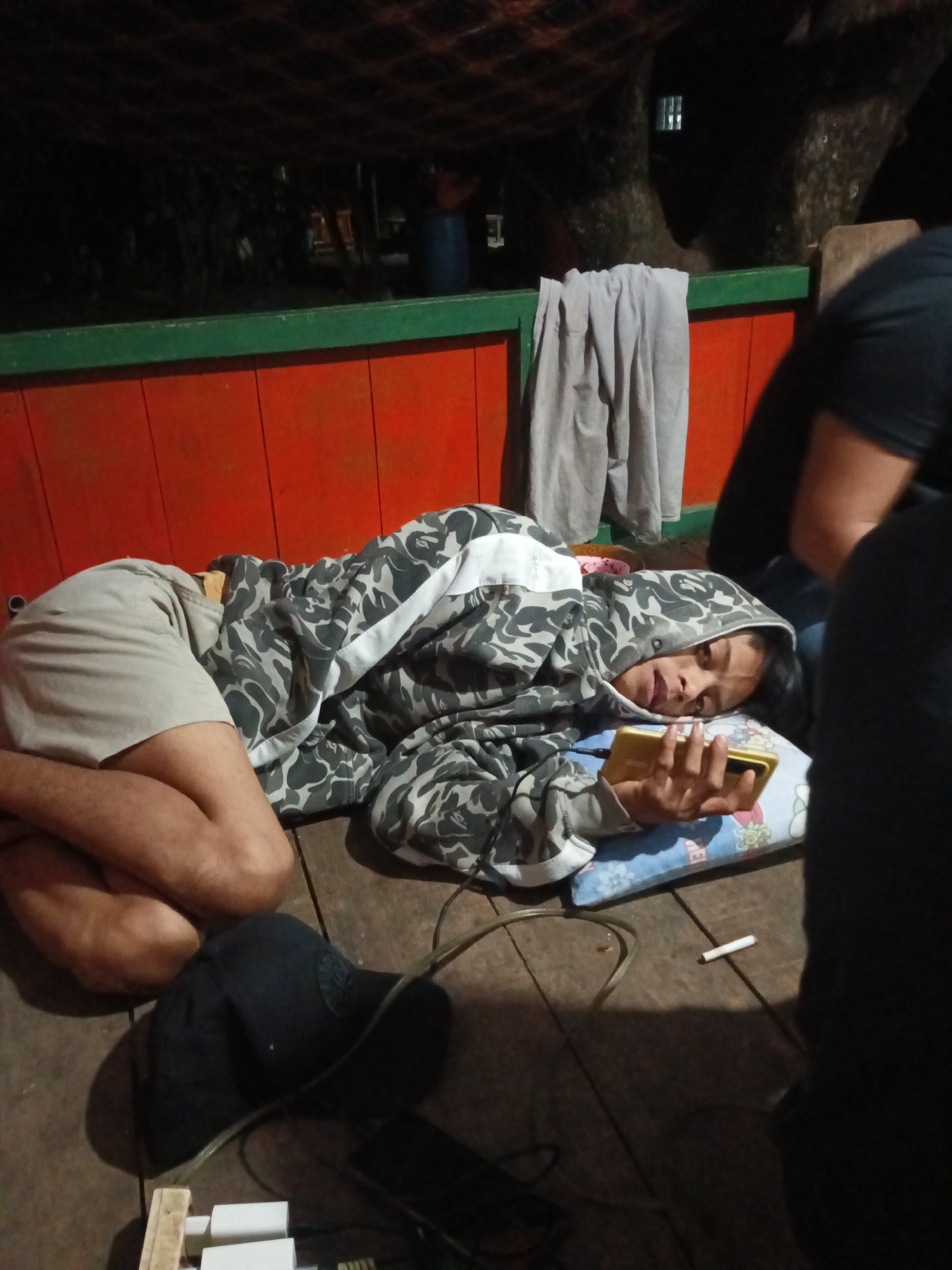In Tumampua, Indonesia, a person is lying on a wooden floor, covered in a camouflage hoodie and shorts, holding a gold smartphone. A blue pillow with a floral pattern supports their head. A lit cigarette rests on the floor nearby. Adjacent to the person is a black baseball cap. In the background, a red and green railing is visible, along with a grey garment hanging on a string and trees. The scene appears to be outdoors, under a roof or netting.