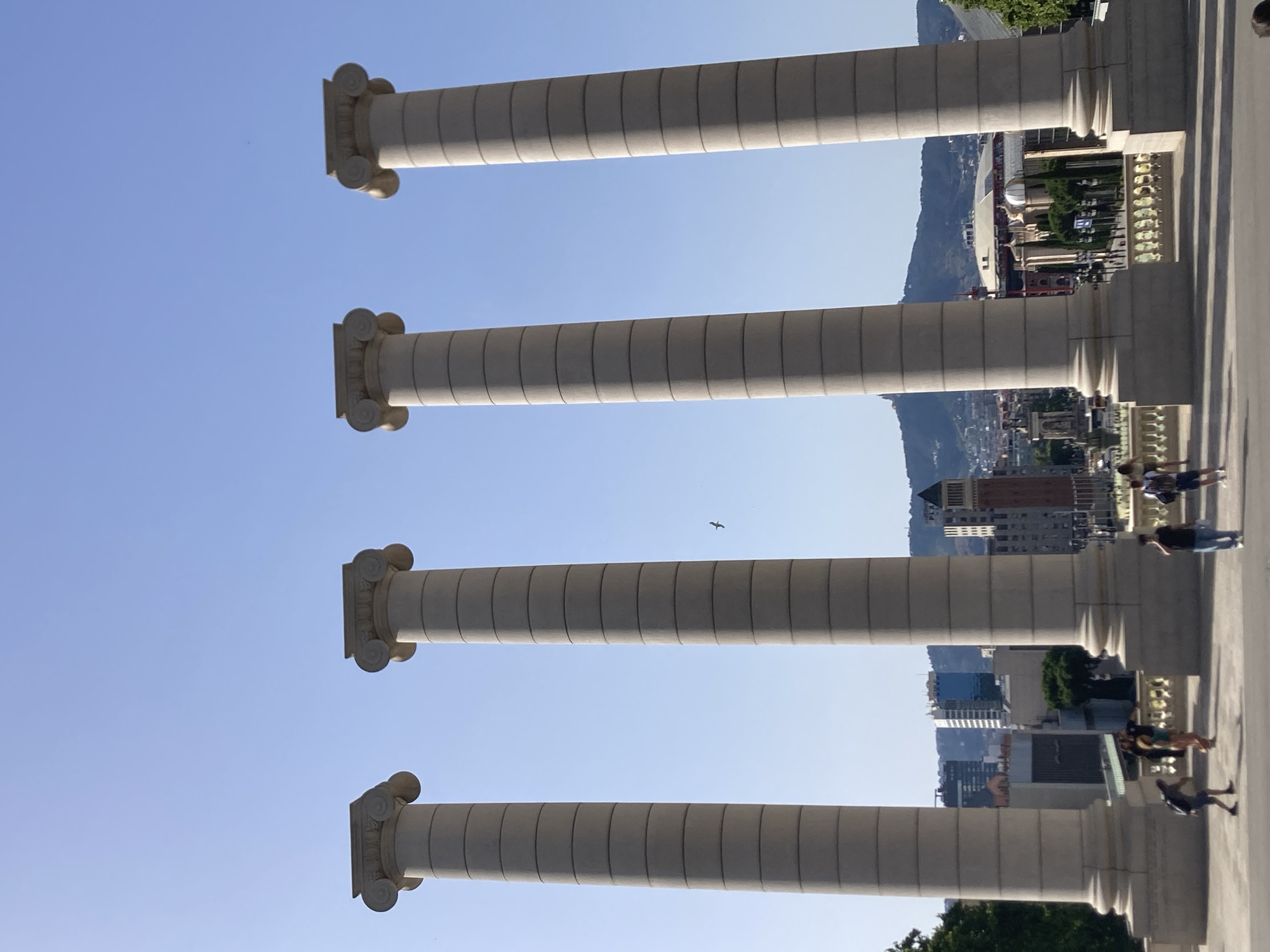 This is a medium shot photograph of the Four Columns of Carles Buïgas in Barcelona, Spain. The columns are tall and made of concrete, with fluted shafts and ornate Corinthian capitals. They are arranged in a line from left to right, with the furthest column on the left partially obscured by a tree. The sky is a clear blue, with a few faint clouds visible. In the background, the city of Barcelona is spread out, with a mix of modern and older buildings. The photo is taken from a slightly elevated position, looking down at the city. A few people are visible in the foreground, walking and taking pictures. The overall impression is one of grandeur and history, with a sense of the vastness of the city.