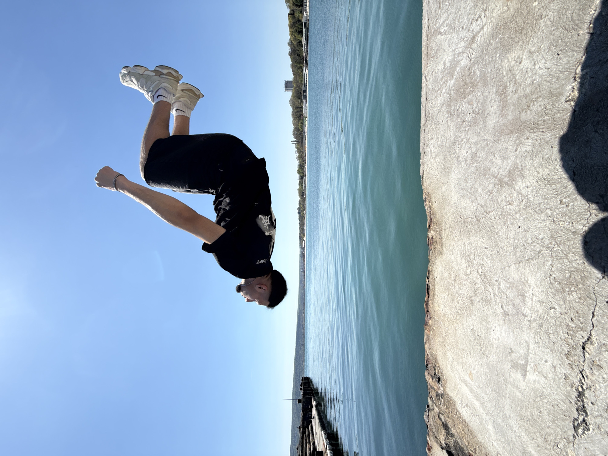 A man is performing a backflip mid-air near the waterfront, specifically near the pier in Varna, Bulgaria. The man is wearing black shorts, a black t-shirt, and white sneakers with black socks. He has a dark hairstyle and appears to be in motion. The background consists of a blue sky on one side, while the other side features the turquoise-colored Black Sea stretching towards the horizon, with a hint of greenery and buildings in the distance. The pier itself is made of concrete and is weathered in appearance. The photo is taken from a low angle, emphasizing the man's height and movement during the flip.