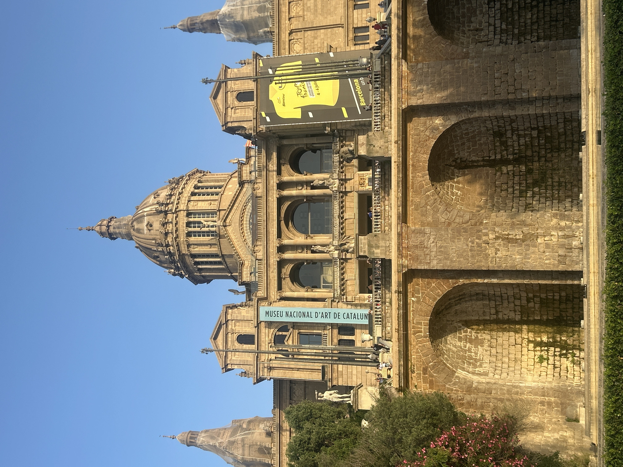 The image shows the Palau Nacional, also known as the National Museum of Art of Catalonia, located on Montjuïc hill in Barcelona, Spain. The grand building features a large central dome, flanked by two smaller domes or towers on either side. The facade is made of stone and exhibits classical architectural elements such as columns and arches. A banner with a yellow jersey design, likely for a sporting event, hangs from the building. Another blue banner displays text in Catalan, "MUSEU NACIONAL D'ART DE CATALUNYA". Several people are visible on a balcony and on the grounds in front of the building, suggesting it is open to the public. The lower section of the image reveals a stone wall with arched openings and a green hedge in the foreground. The sky is clear and blue, indicating fair weather.