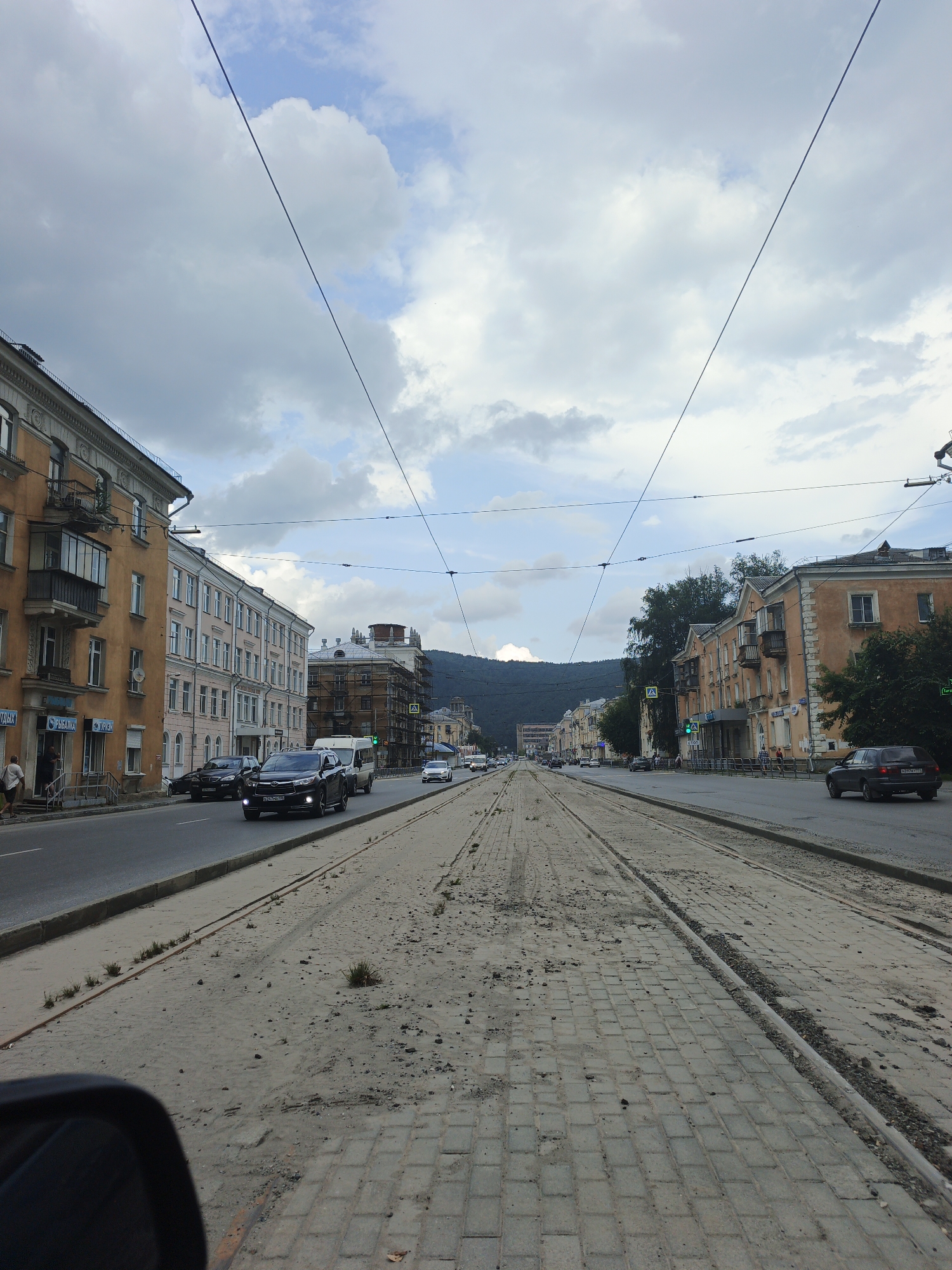 The image captures a street scene in Zlatoust, Russia, likely depicting an urban environment with a tram line under construction or repair. The perspective is from a vehicle, looking down a wide, straight street. On the left, a multi-story building with an orange facade features balconies and arched windows. Adjacent to it, a lighter-colored building has a similar architectural style. Across the street, on the right, are more buildings, some with orange exteriors and prominent rooflines, and a green tree line separates the buildings from the street.
The foreground is dominated by a tram track embedded in a sandy, rough surface, with cobblestones visible on the right side of the street. The tram tracks appear to be either under construction or disused, with debris scattered around them. Several vehicles are on the road, including a black SUV in the middle lane and other cars in the distance. The sky is overcast with a mix of white and gray clouds, and overhead, power lines stretch across the frame. In the background, hills or mountains are visible, partially obscured by clouds. The overall impression is that of a functional but somewhat weathered urban setting.