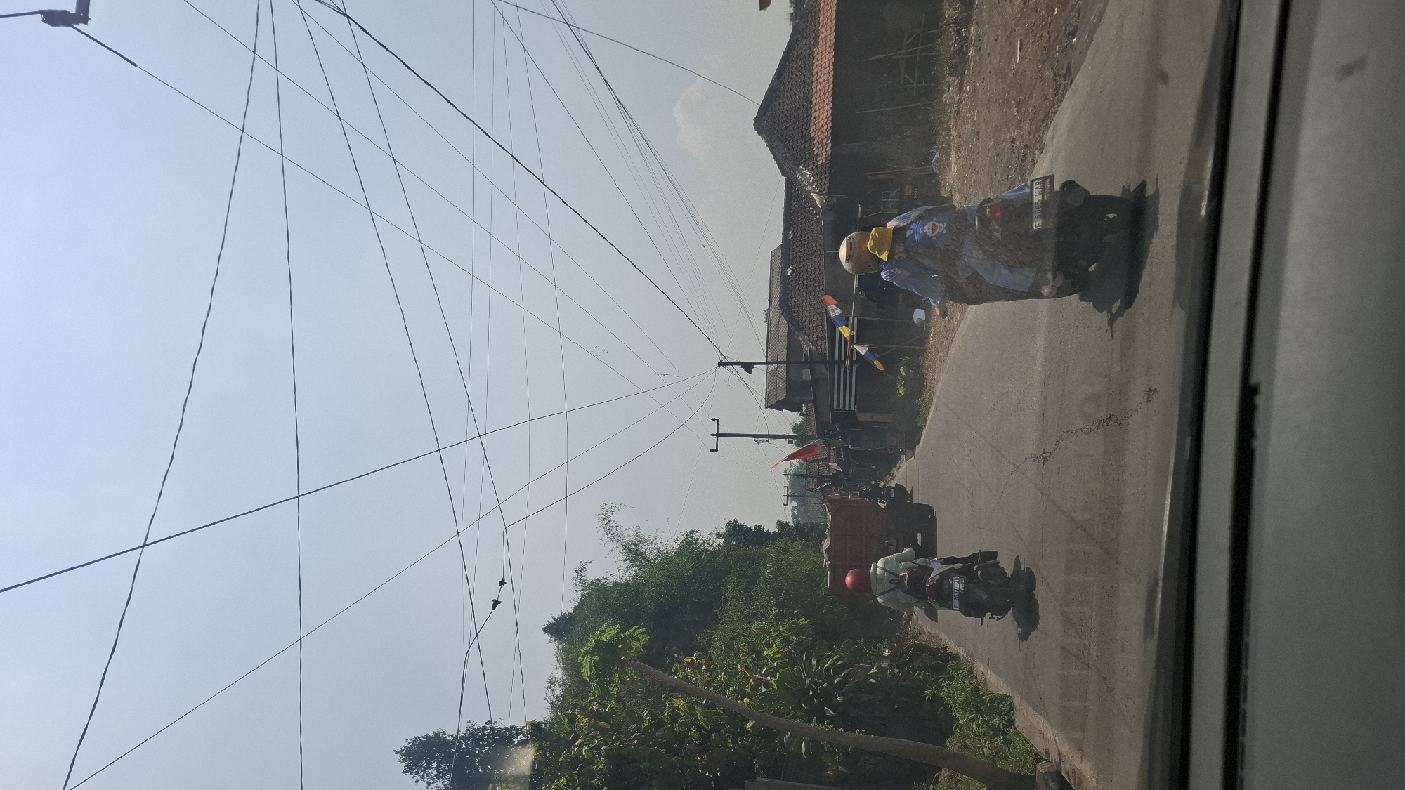 This image captures a street scene in Sirnagalih, Indonesia, under a clear, bright sky. Multiple power lines crisscross the sky, creating a dense network above the road. The road itself is asphalted and appears somewhat worn, with visible cracks.
In the foreground, the interior of a vehicle is partially visible, suggesting the photo was taken from inside a car. On the road, two motorcycles are in motion. The motorcycle in the left lane is ridden by a person wearing a red helmet and a light-colored jacket. The motorcycle in the right lane carries two individuals; the rider wears a yellow helmet and the passenger has a yellow hat and a blue backpack with a cartoon character on it.
Further down the road, a large, orange truck is visible, also in motion. To the right of the road, several houses with traditional tiled roofs are situated. Red and white striped flags are displayed near some of the houses, possibly indicating a celebration or national holiday. Dense green foliage lines the left side of the road, including trees and bamboo. The overall impression is of a typical day on a rural or semi-urban road in Indonesia.