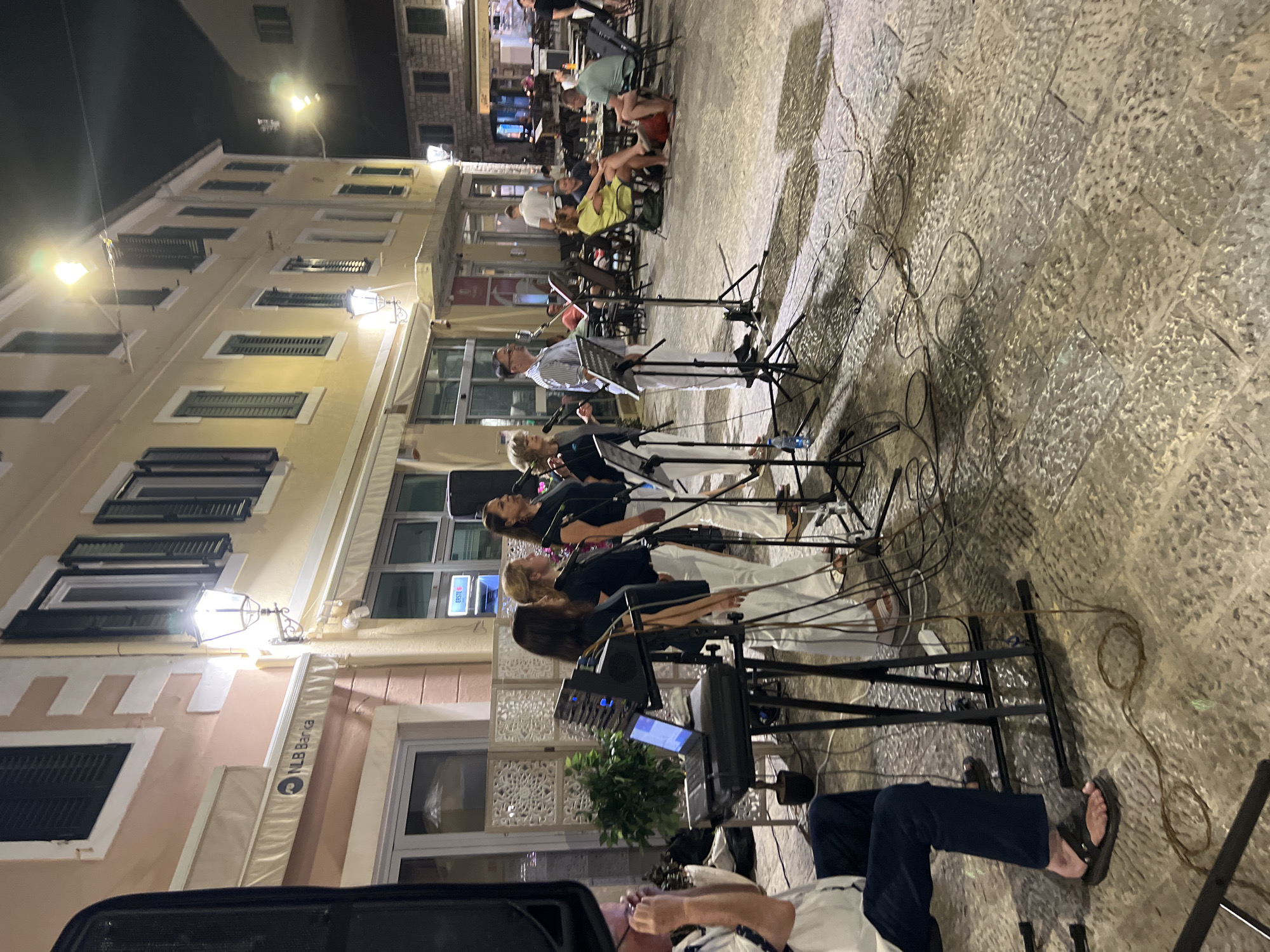 In Herceg Novi, Montenegro, a group of five singers, four women and one man, are performing on a cobblestone street in front of buildings. The singers are dressed in white pants and dark tops, and they are holding microphones. They are standing behind music stands with sheet music. A man in a white shirt and dark pants is seated to the left, possibly operating a laptop on a keyboard stand. The background shows a building with awnings and windows, a street lamp, and an outdoor seating area with people dining and socializing. The scene appears to be an evening performance, with streetlights illuminating the area.