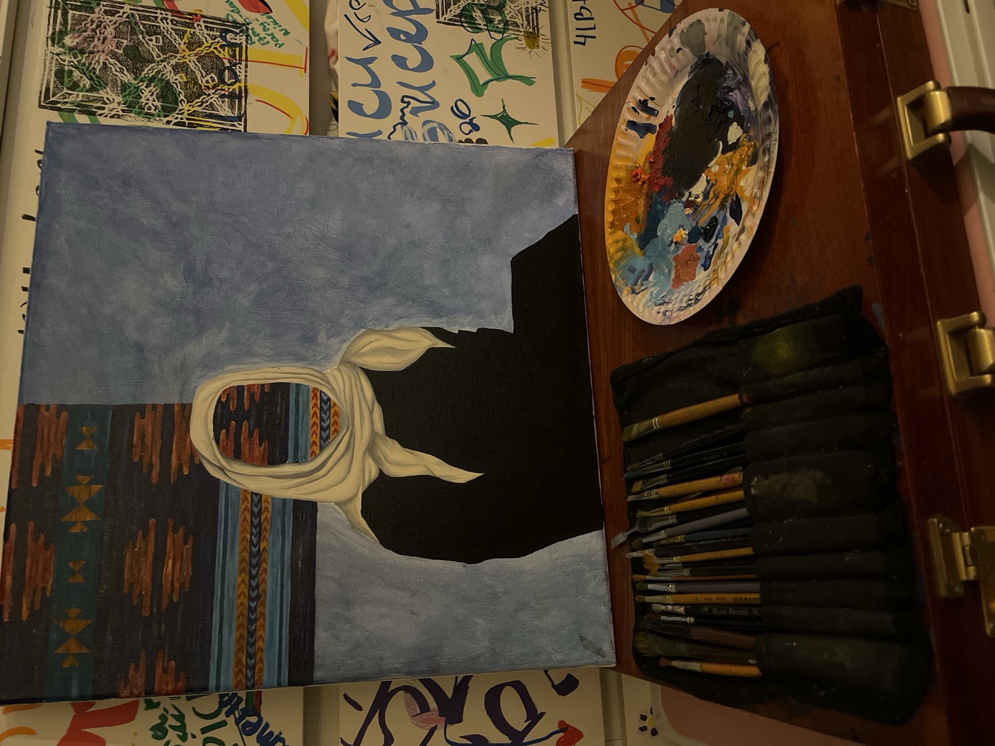 The image shows an unfinished painting on a canvas. The painting depicts a figure wearing a headscarf, with the background partially filled in with blue sky. The lower portion of the painting features a patterned textile. The right side of the image shows a wooden box with a handle and a brush holder with many brushes inside. A paint palette with used paints is above the brushes. The painting and the painting materials are placed on top of multiple canvases.
There is no evidence of the city of London, United Kingdom.