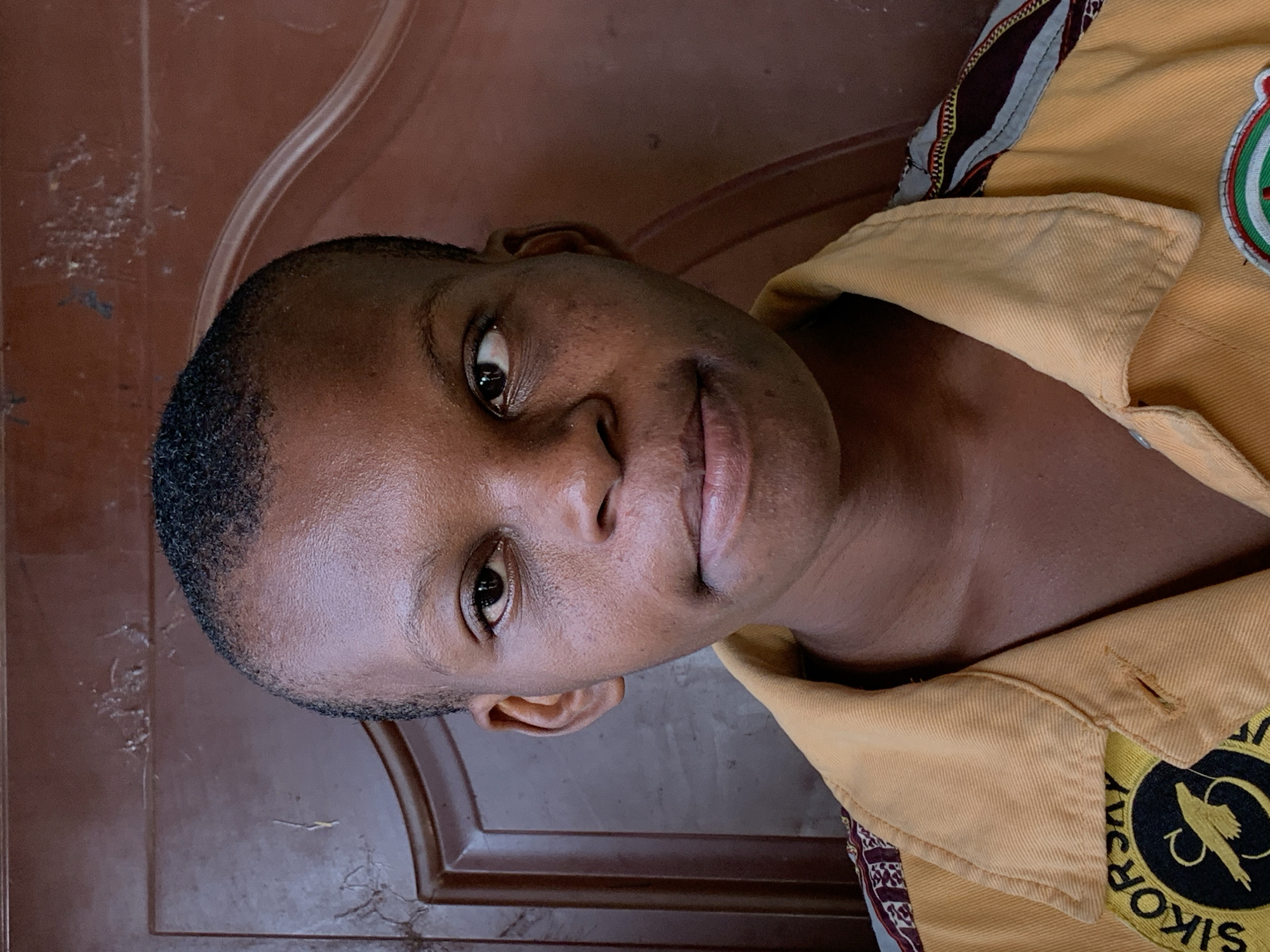 A close-up, rotated portrait shows a dark-skinned person with short, dark hair. The individual is wearing a yellow collared shirt with patches, one of which reads "SIKOR", possibly referring to a location or organization. The background appears to be a brown, possibly wooden, surface. The location is potentially in Ikorodu, Nigeria, given the text on the garment.