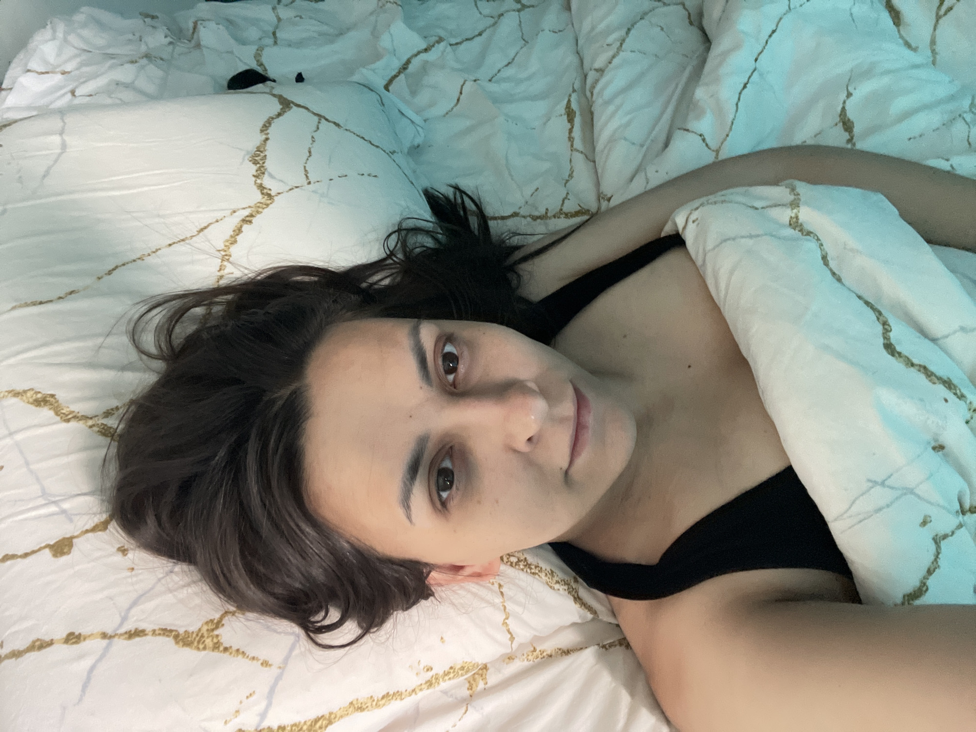 A single individual is depicted in a close-up, high-angle photograph, lying supine on a bed. The subject has dark brown hair, dark brown eyes, a visible nose, and lips with a slight upturn, suggesting a subtle smile. A small mole is discernible above the upper lip. The subject is wearing a black, thin-strapped garment on the upper torso. The bed is furnished with white bedding that features a distinct pattern of irregular, thin, gold-colored lines, reminiscent of marble veining. A pillow with the same pattern supports the subject's head, and a duvet or blanket, also patterned, partially covers the subject's torso. The scene is illuminated with a cool, slightly bluish-green light. The location is an unknown interior setting within the city of Unknown, Unknown.