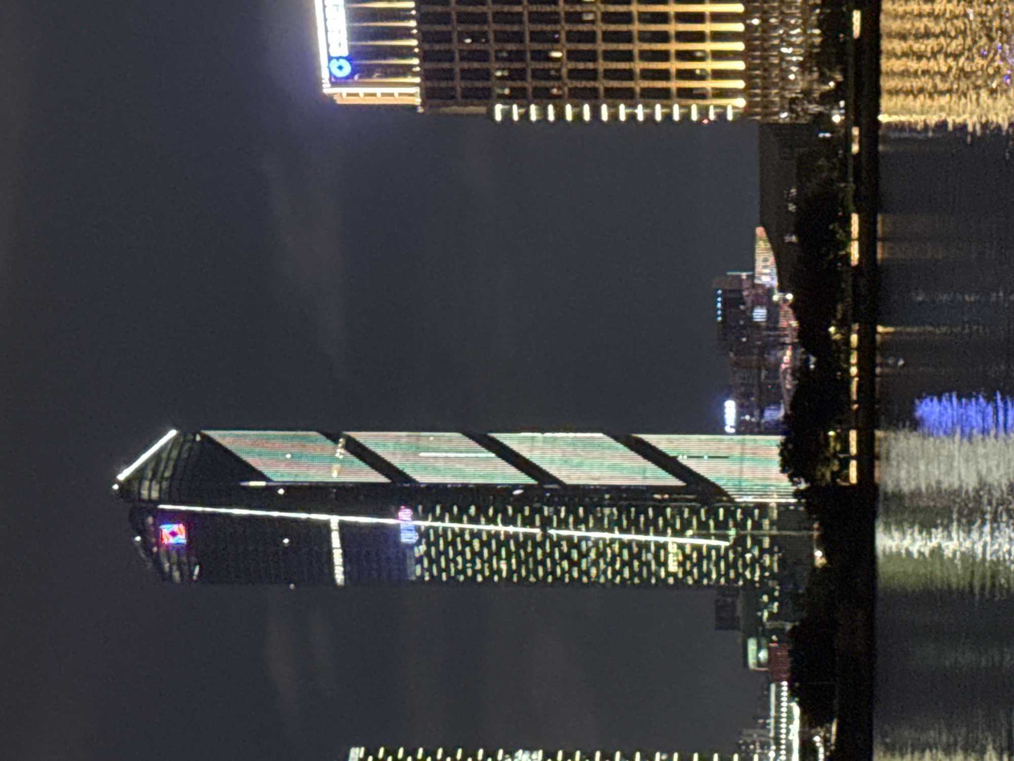 This forensic description details a nighttime cityscape in the Tianhe District of Guangzhou, China, showcasing three prominent illuminated skyscrapers reflected in a body of water, likely the Pearl River.
The central and most dominant structure is the **Guangzhou CTF Finance Centre** (East Tower), a very tall, slender skyscraper. Its dark facade is strikingly illuminated with vertical white light strips and a distinct pattern of diagonal, segmented light bands that display a dynamic array of colors, primarily green and pink/red. The building culminates in a sharp, angular, brightly lit white crown. Below the crown, a square, glowing red logo with a white symbol is visible, and further down, another illuminated sign appears to display "GUANG ZHOU".
To the right of the central skyscraper stands another tall building, likely the **Rosewood Guangzhou**. This structure is characterized by warm, golden-yellow vertical lights illuminating its upper floors and facade, giving it a grand appearance. At its apex, a prominent blue "C" logo is visible, followed by additional illuminated text.
On the far left, a third skyscraper, likely the **Guangzhou International Finance Center** (West Tower), is partially visible. It is also illuminated with numerous white lights, appearing as a grid of horizontal and vertical elements.
A dark, narrow strip of land, dense with trees, separates the illuminated buildings from the foreground water. The water, calm and dark, strongly reflects the vibrant lights of the skyscrapers, creating undulating patterns of white, gold, and blue. The night sky above is a dark grey, indicating cloud cover. No people, specific objects, or discernible actions are present in the scene.