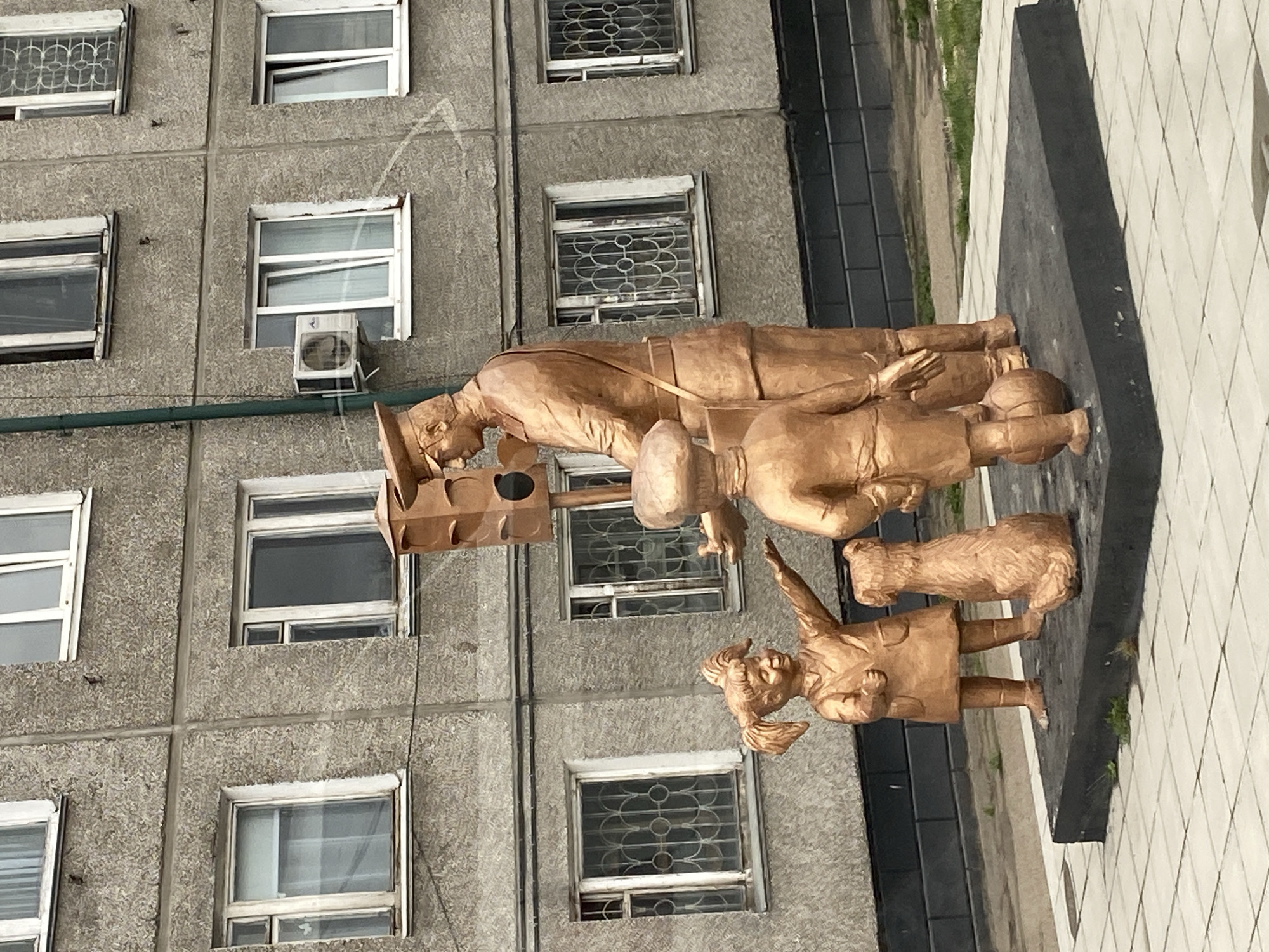 A statue in the city of Usolye-Sibirskoye, Russia, shows a uniformed postman with a mailbag walking with two children and a dog. The statue stands in front of a gray building with multiple windows.