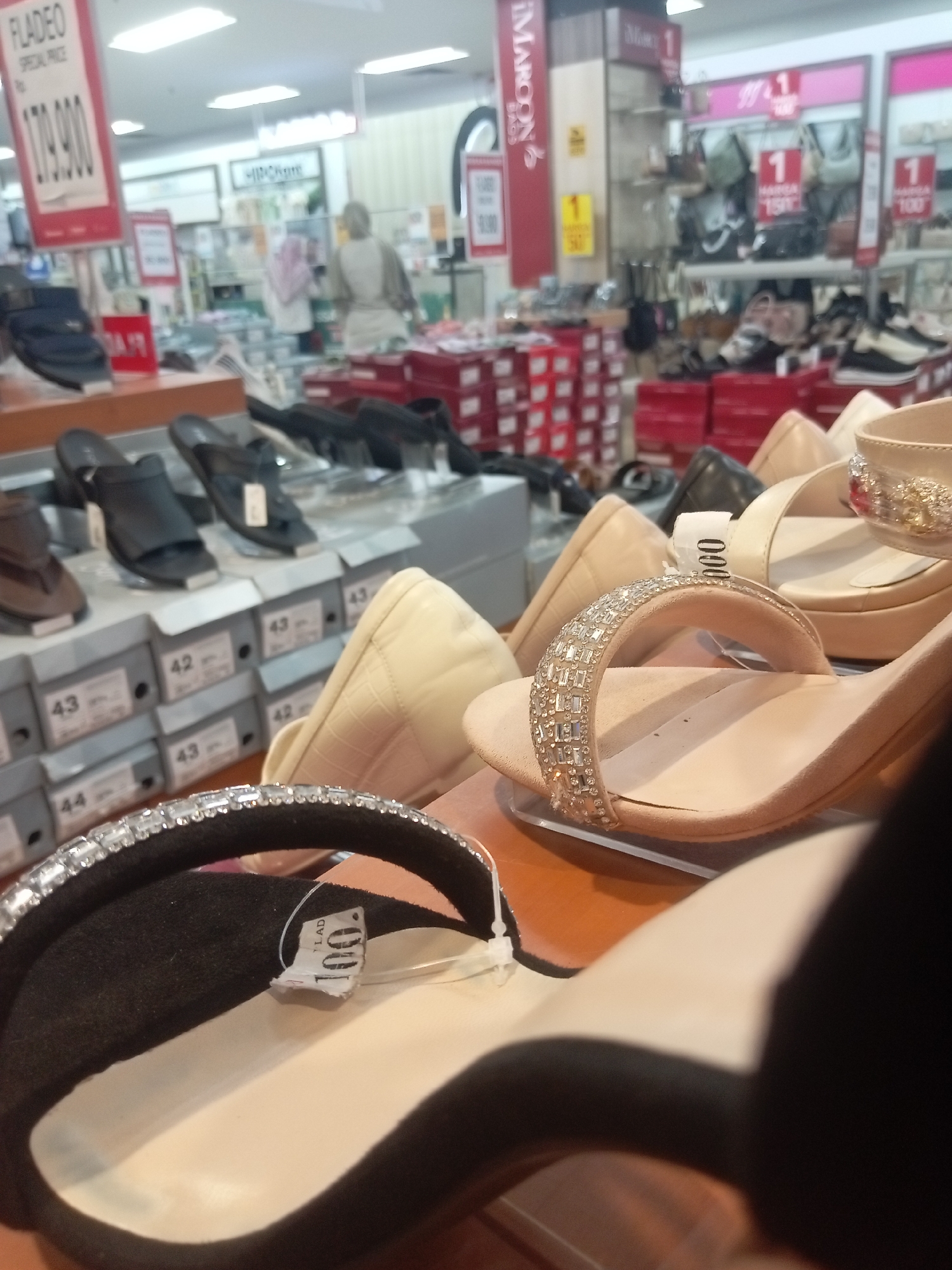 The image shows a retail store in Batam, Indonesia, likely a shoe store. In the foreground, there are several pairs of women's shoes on display. One pair of beige high-heeled sandals with rhinestone embellishments on the strap is prominent. Another black sandal with a similar rhinestone-decorated strap is partially visible. In the background, shelves are stocked with more shoes and shoe boxes. Price tags and promotional signs are visible, including one with "FLADEO SPECIAL PRICE RP. 179.900" and another with "100." Behind the displays, a woman in a light-colored garment is seen interacting with merchandise. The environment appears to be a well-lit indoor shopping space with typical retail fixtures.