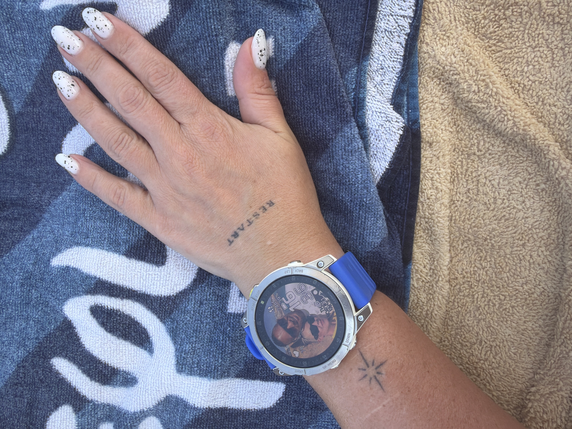 The image shows a close-up of a person's wrist wearing a Garmin watch. The watch face displays a photo of two people, the time "16:36," and various fitness metrics. The wrist has two tattoos: a small starburst and the word "RESTART" written vertically. The person's hand, with white nail polish speckled with black dots, is visible in the upper right portion of the image. The background consists of a textured blue and white towel, likely a beach towel, placed on a tan-colored terry cloth surface. The context suggests an outdoor setting, possibly a beach or poolside, in Sharm El Sheikh, Egypt.
